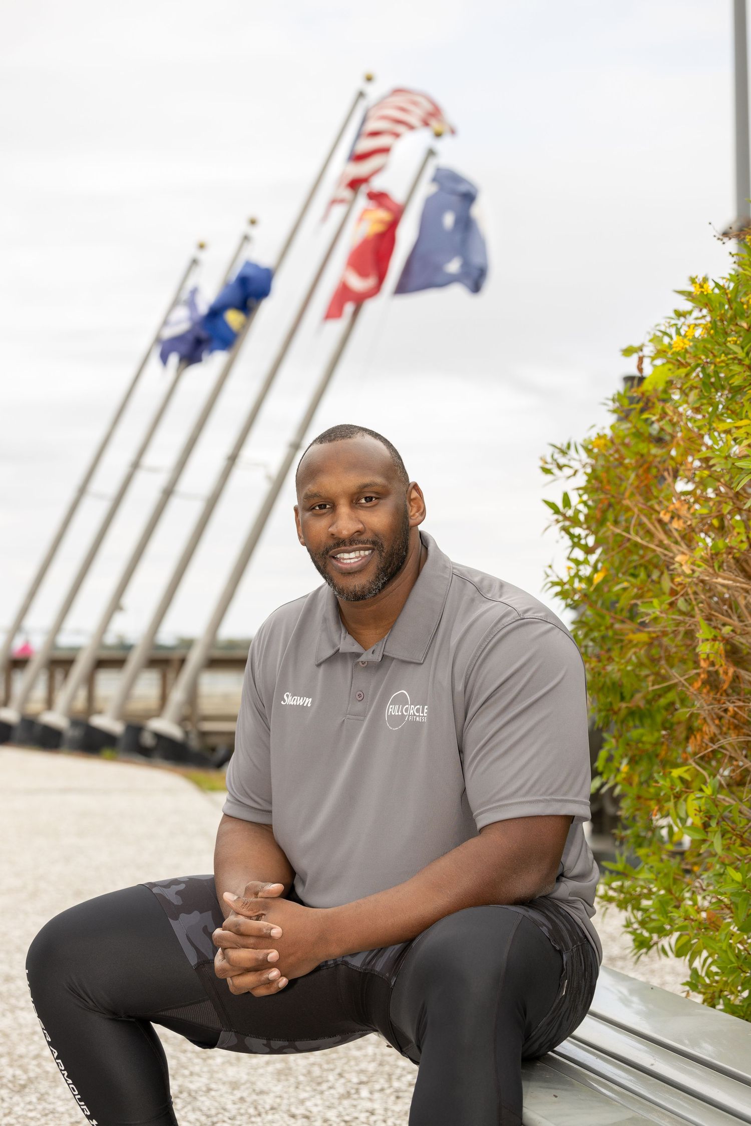 Person in grey polo shirt poses casually near waterfront with flags and autumn scenery.
