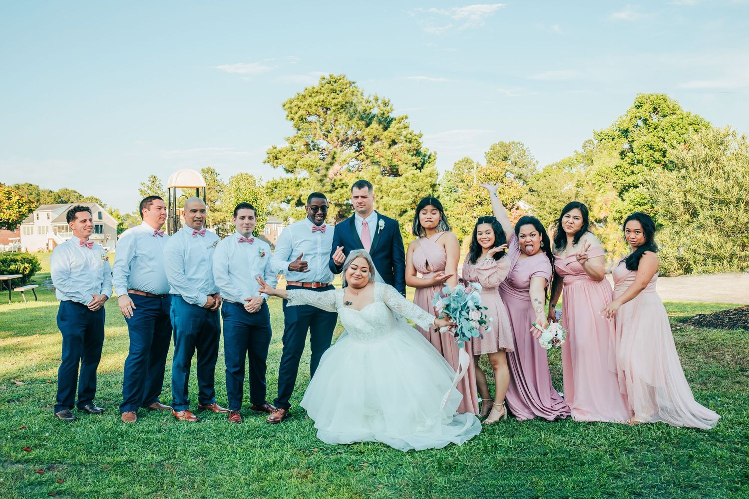 Wedding party in pink dresses and blue suits pose for group photos on grassy lawn.
