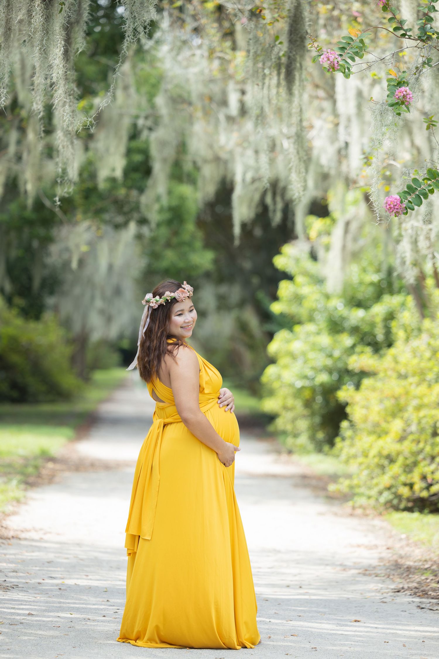 A woman in a flowing yellow dress poses gracefully on a path lined with Spanish moss trees.