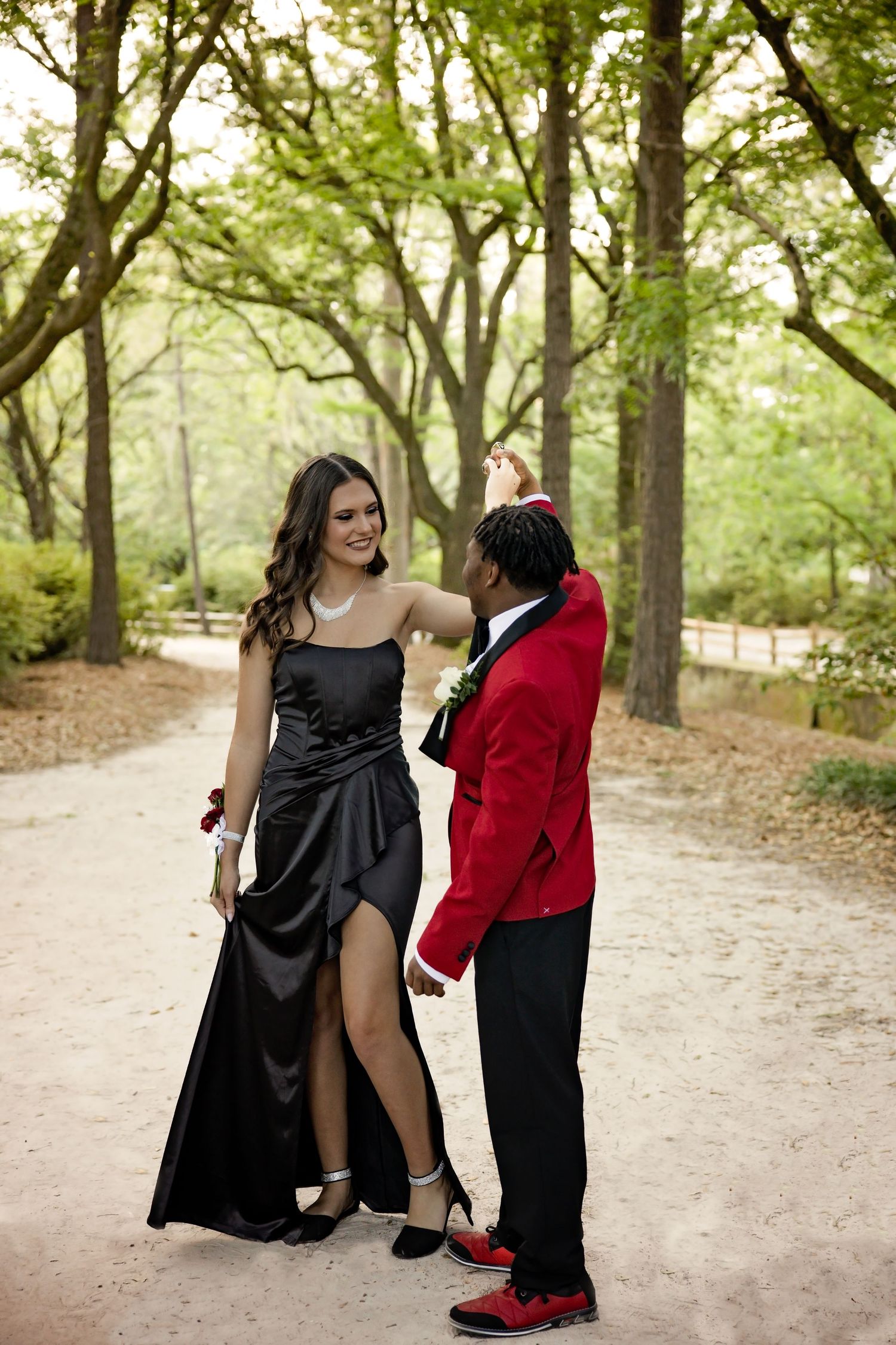 Two people in formal wear pose playfully on a dirt path surrounded by trees.