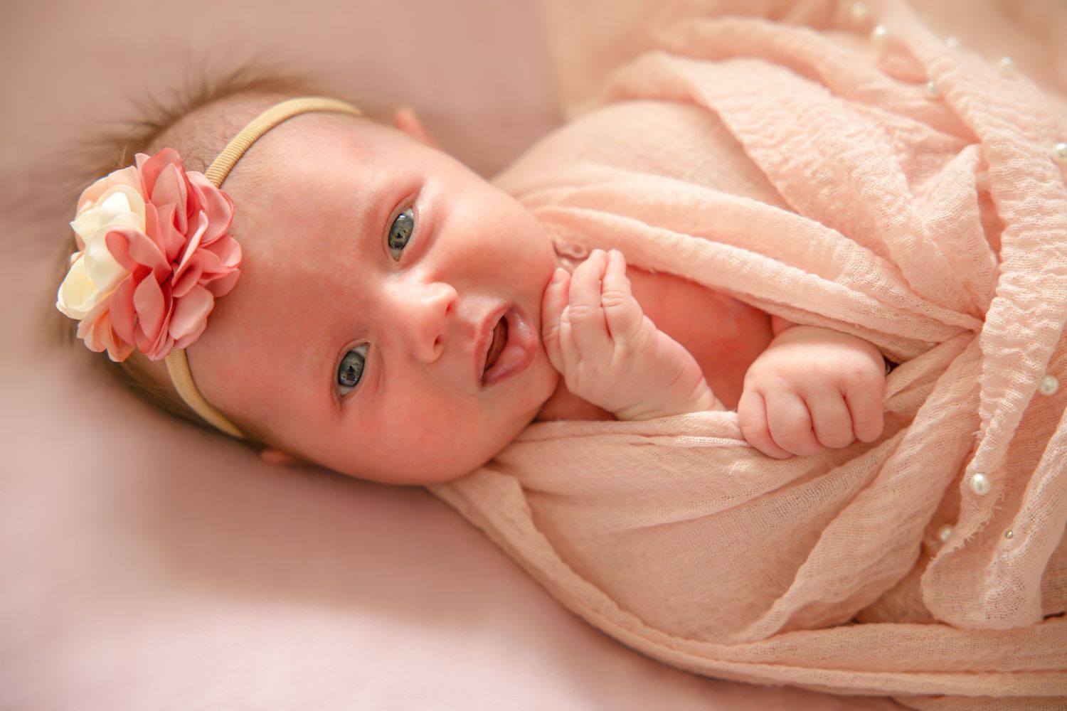 Baby in pink outfit with floral headband lying on soft bedding.