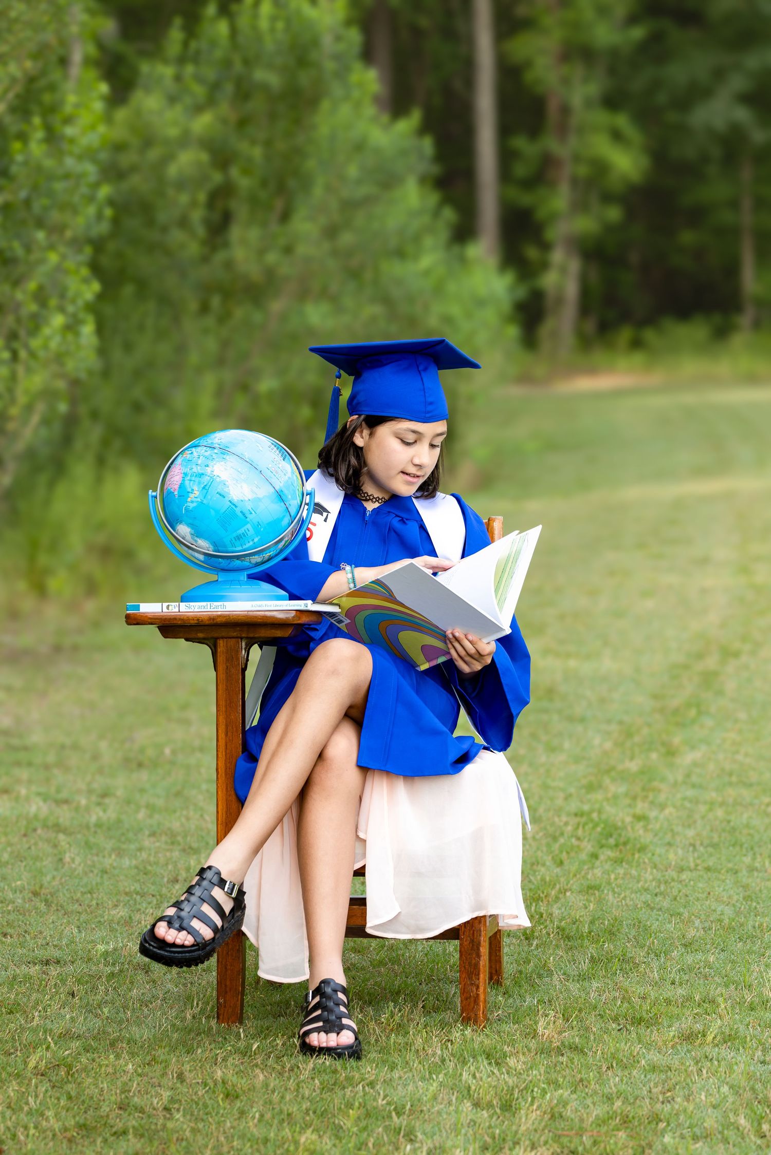 Child in graduation cap reads book while sitting with globe on wooden chair outdoors.