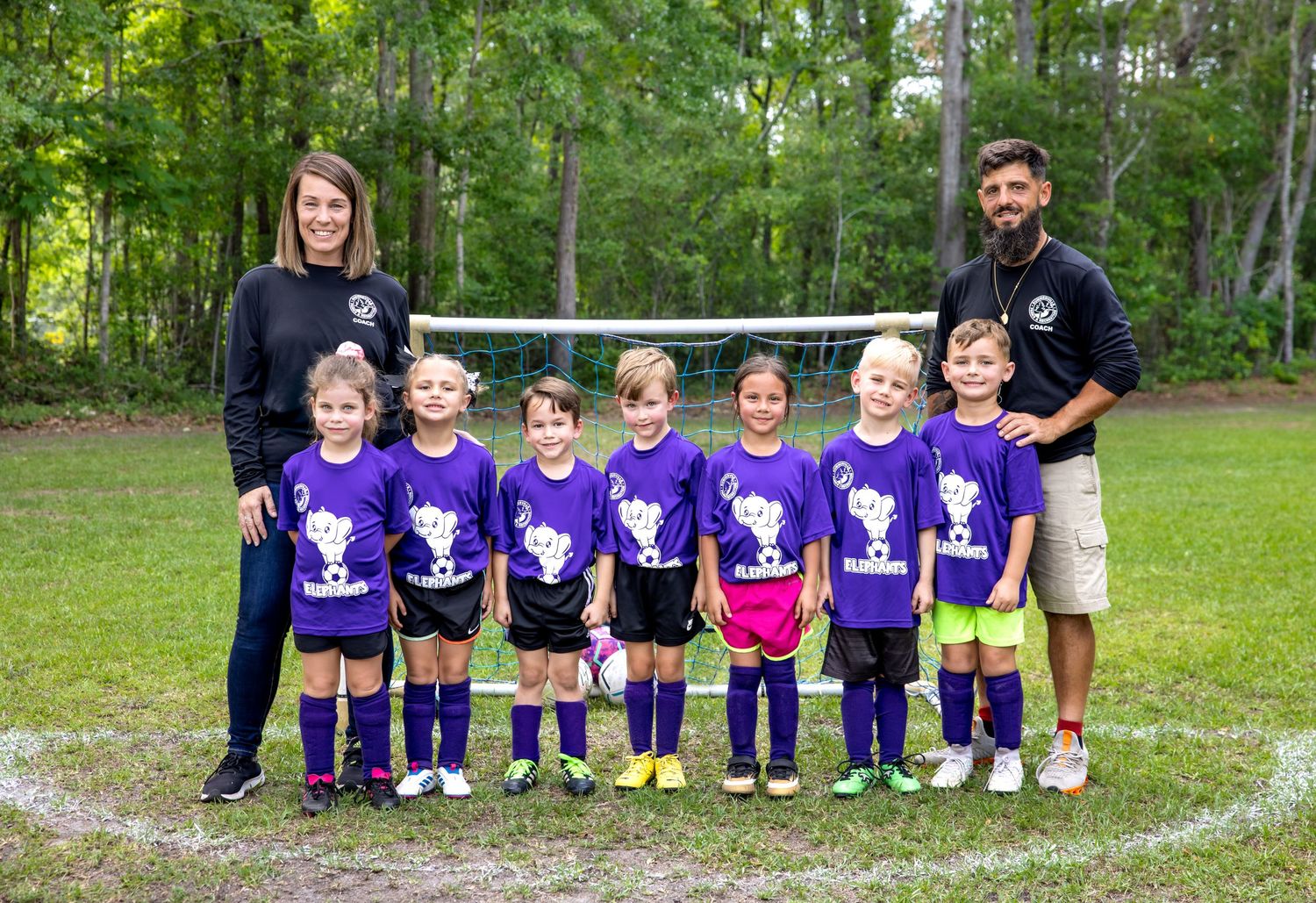 Youth soccer team in purple uniforms posing together in front of trees.