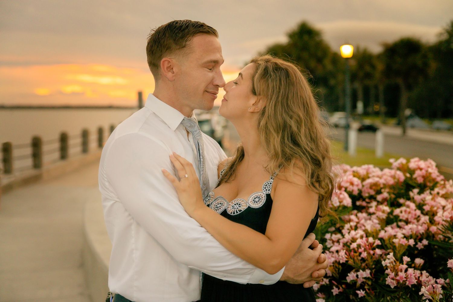 Romantic sunset photoshoot on boardwalk with pink flowers and dramatic orange sky.