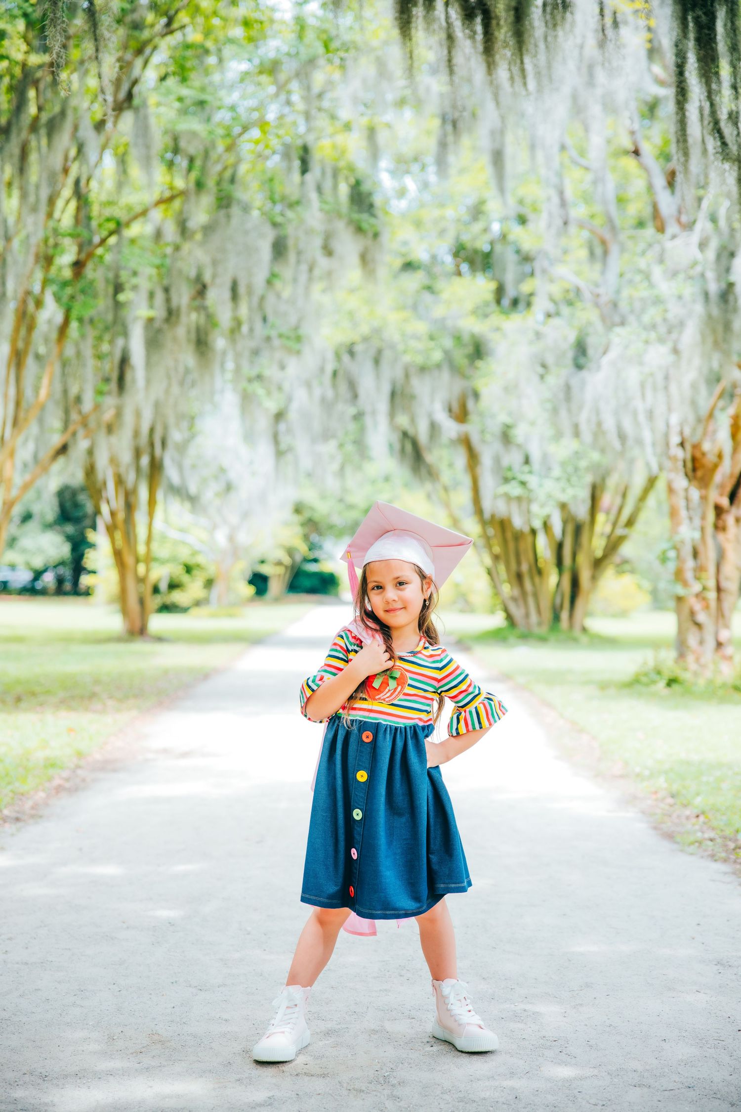 A young graduate in blue and pink outfit strikes playful poses on a park path.