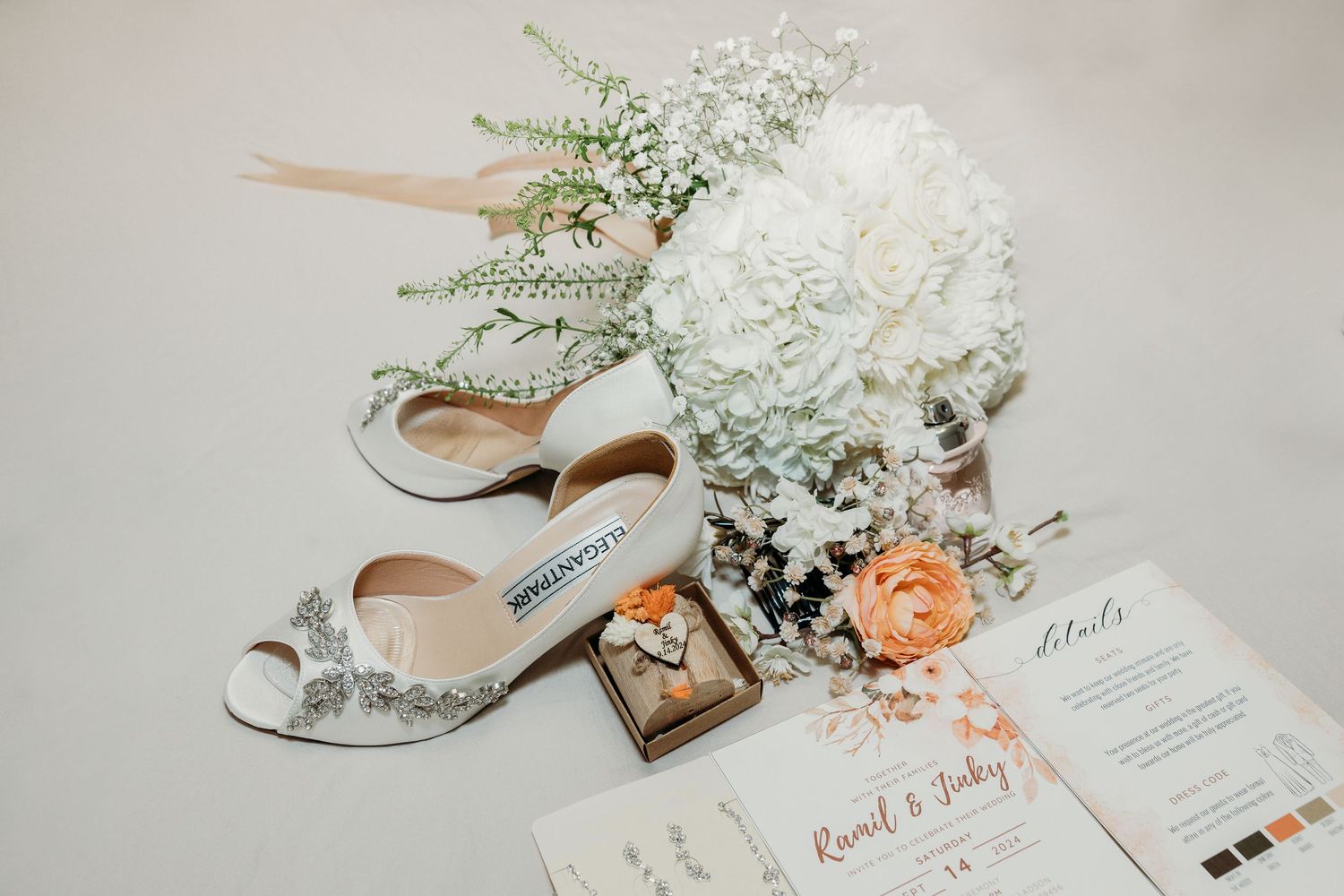 Wedding details including white flowers, silver shoes, and invitation cards arranged on light gray surface.