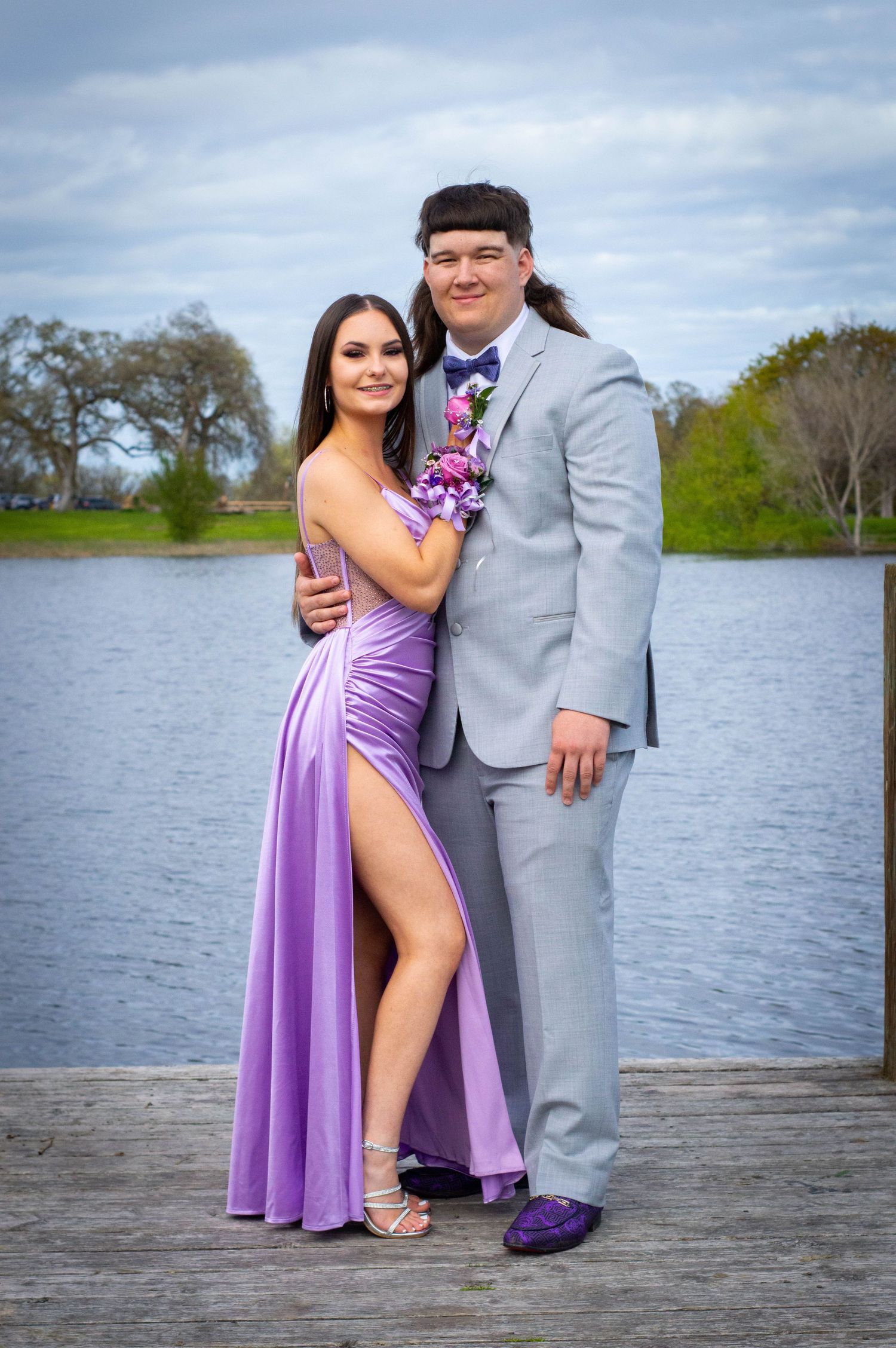 Couple in formal attire standing on wooden dock by water.