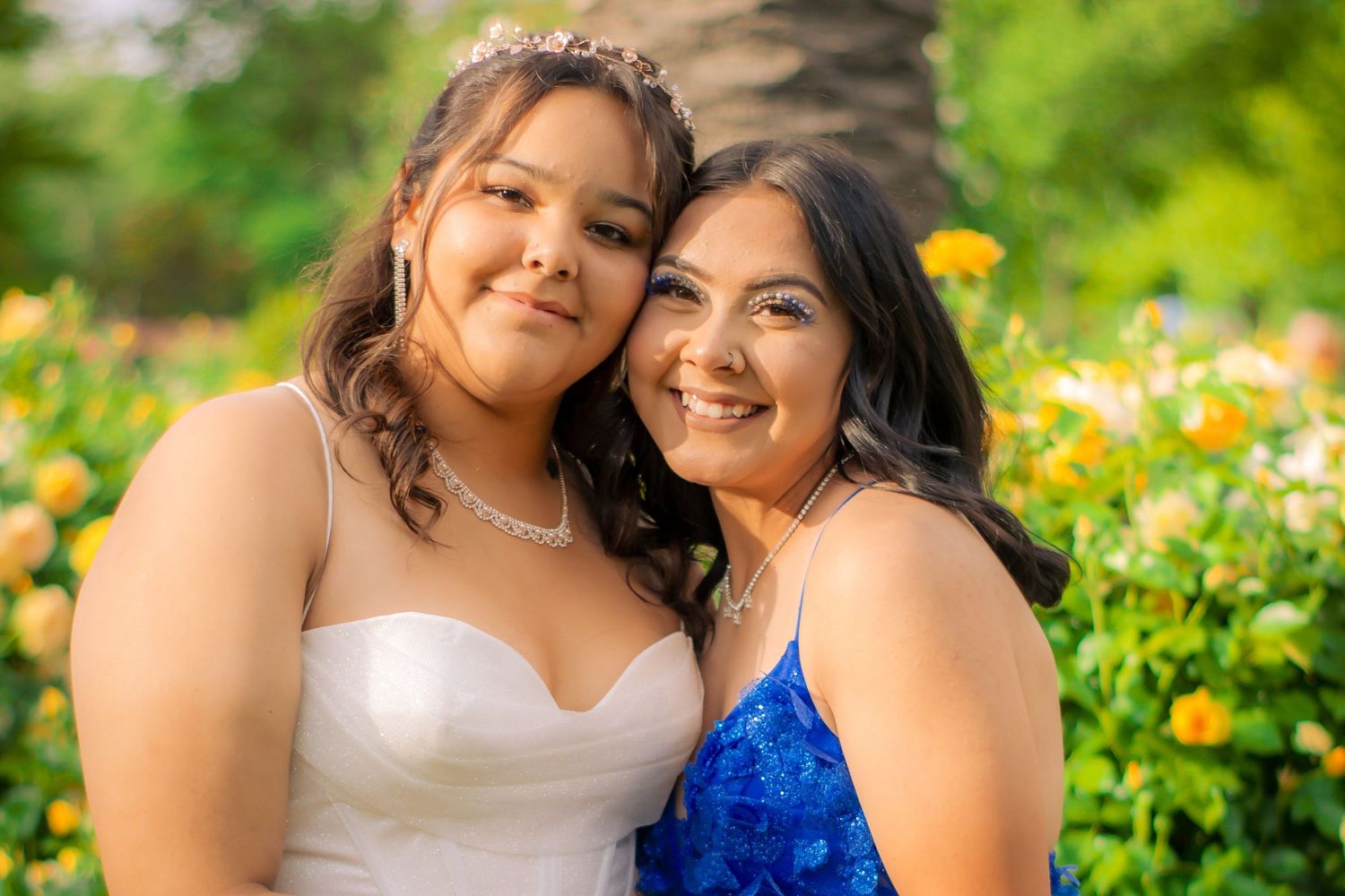 Two friends in formal attire smile together in a garden filled with yellow flowers.
