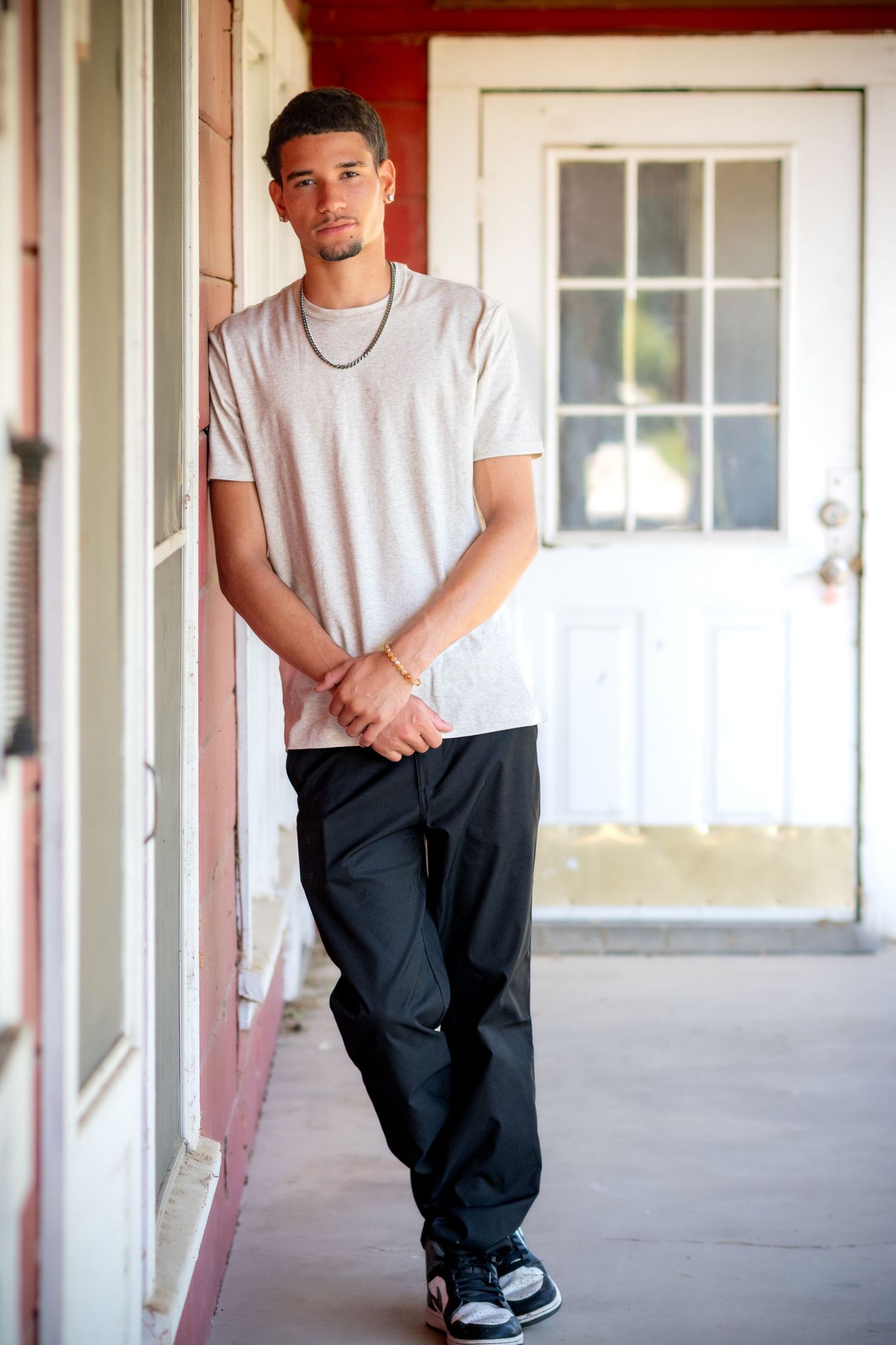 Young man in white t-shirt and black pants leaning against a red pillar on a porch near a white door.