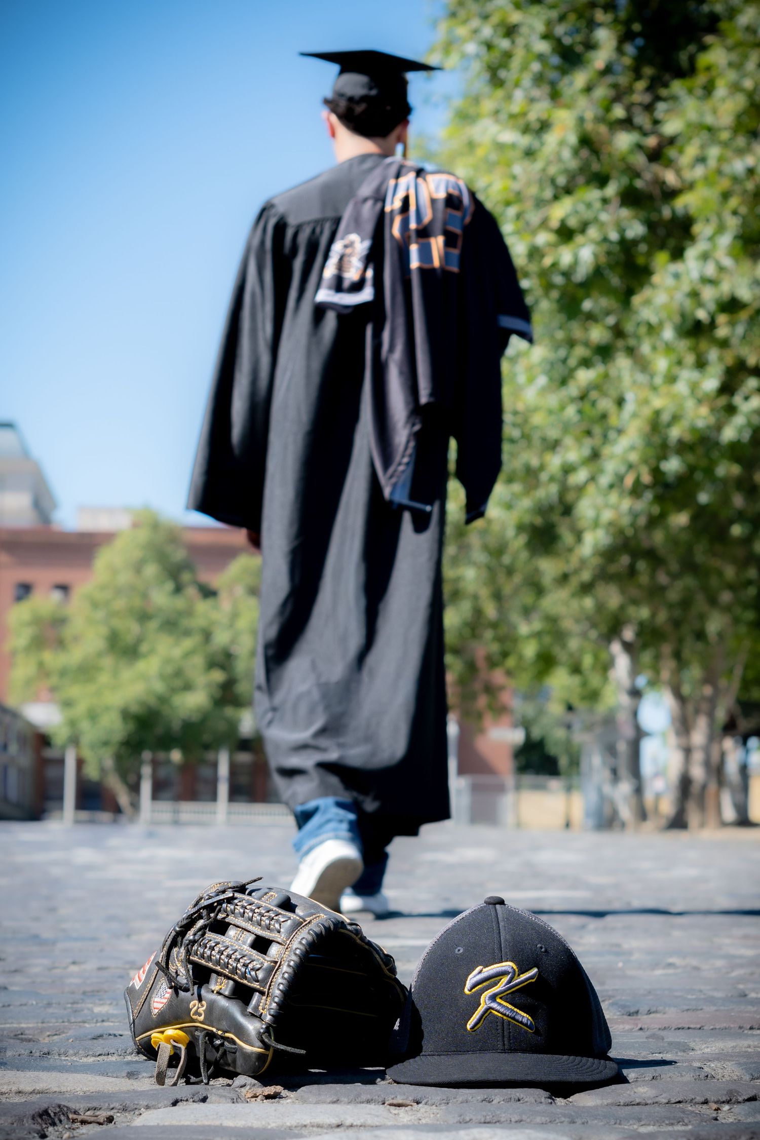 Graduate in black cap and gown walks away, with baseball glove and Rawlings cap in foreground on cobblestones.
