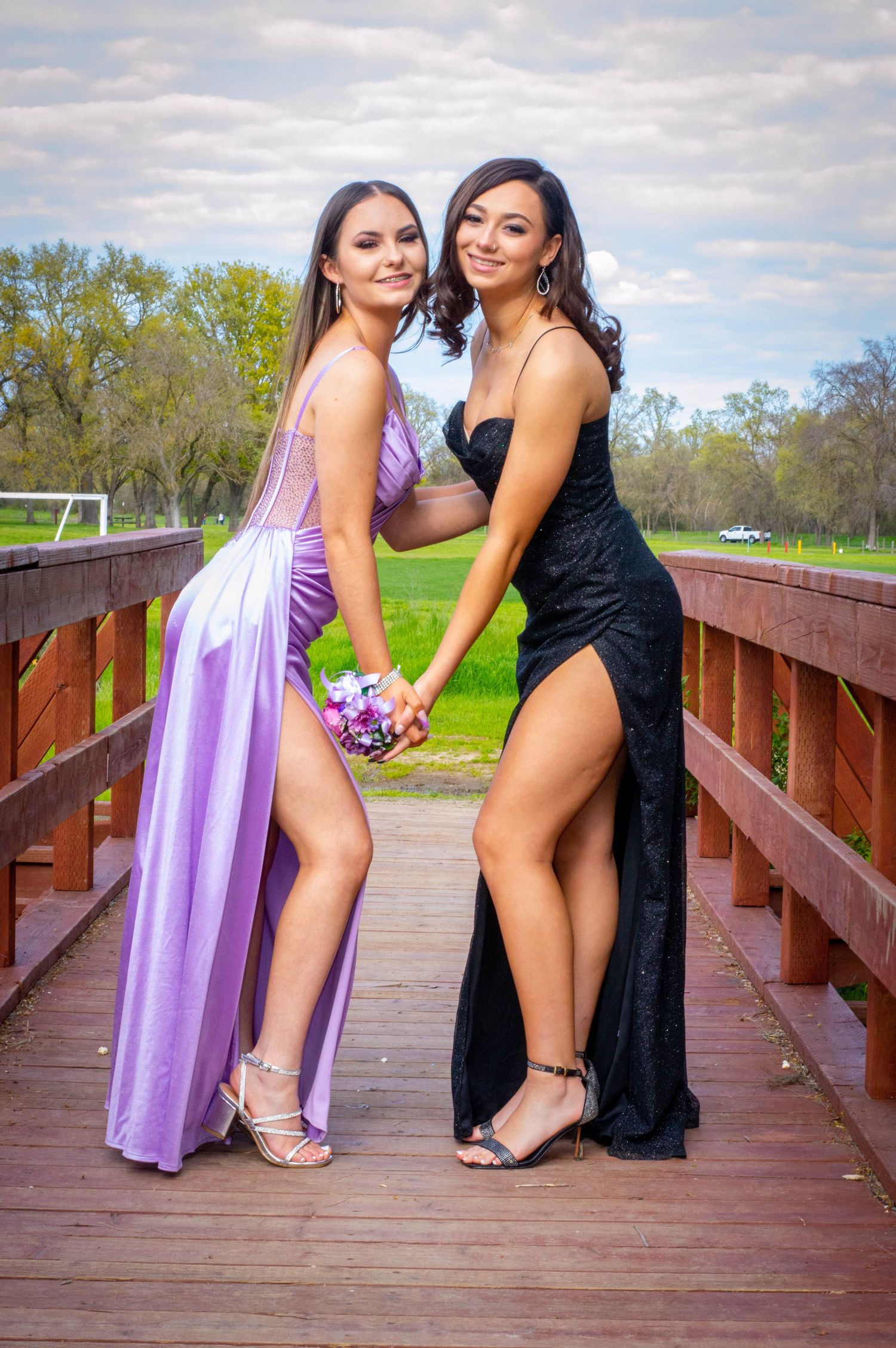 Friends in formal dresses striking playful poses on wooden bridge.