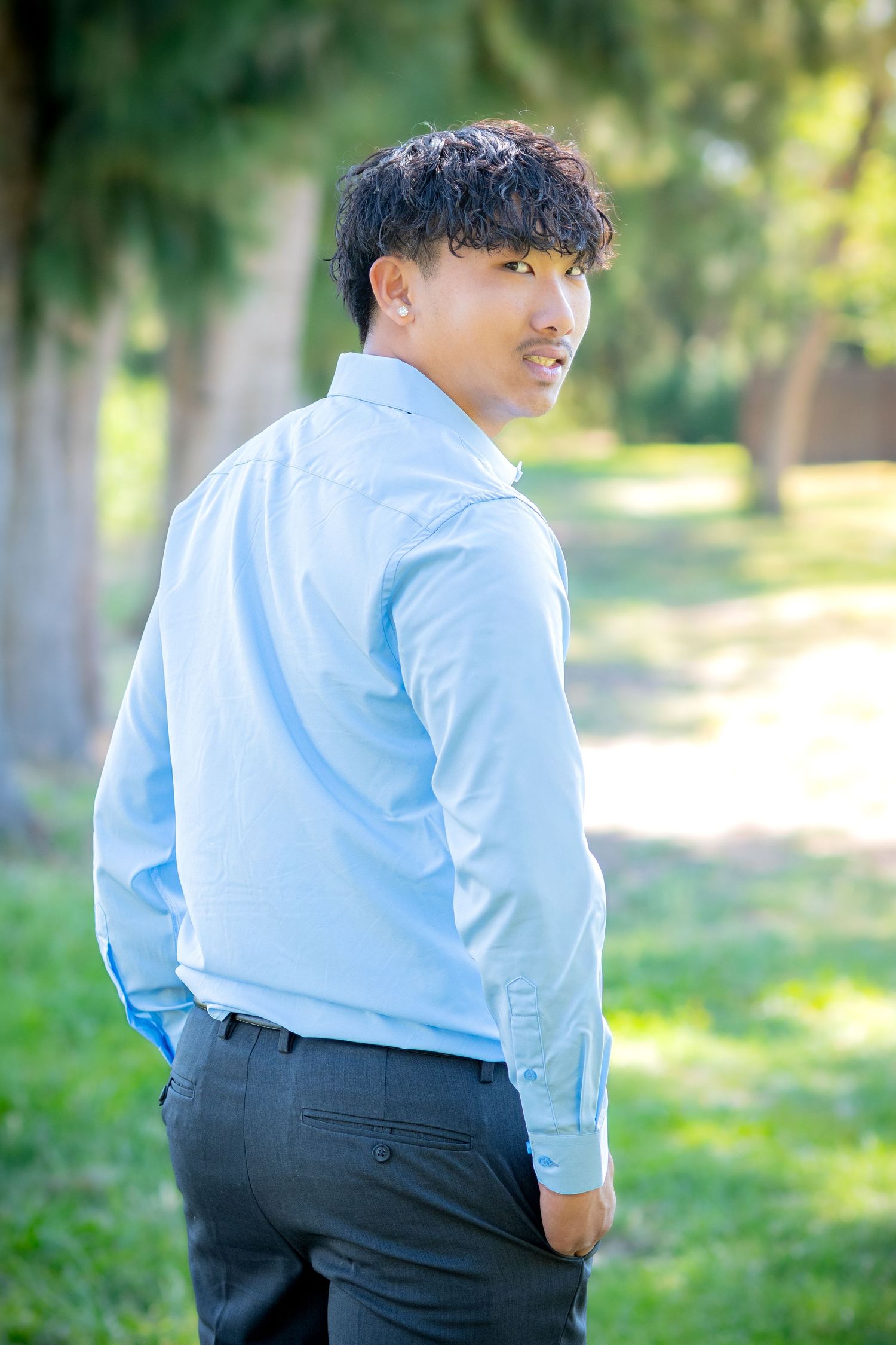 Young man in light blue dress shirt and gray pants glances back over shoulder outdoors in green park setting.