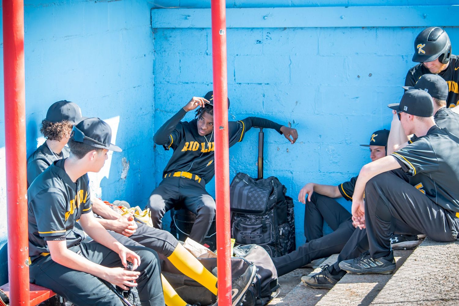 Baseball team dugout with players sitting on bench against blue wall with red posts.