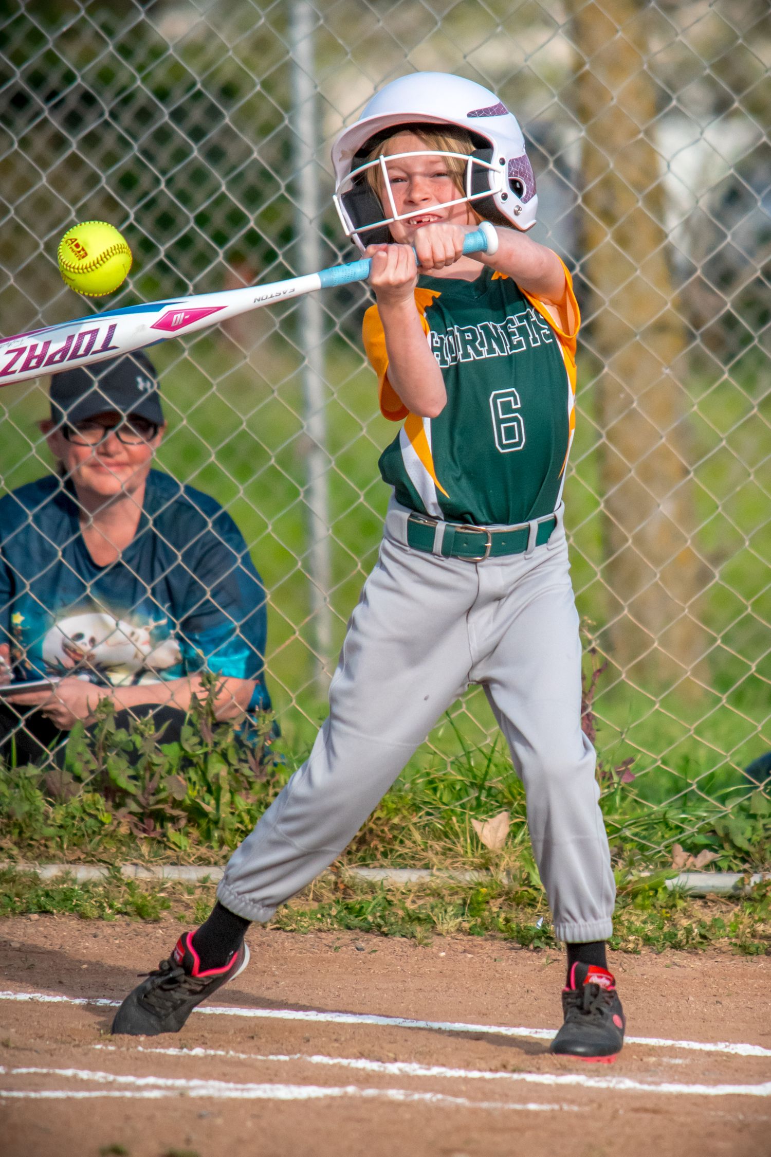 Youth softball player at bat wearing green uniform and helmet, preparing to hit yellow softball.
