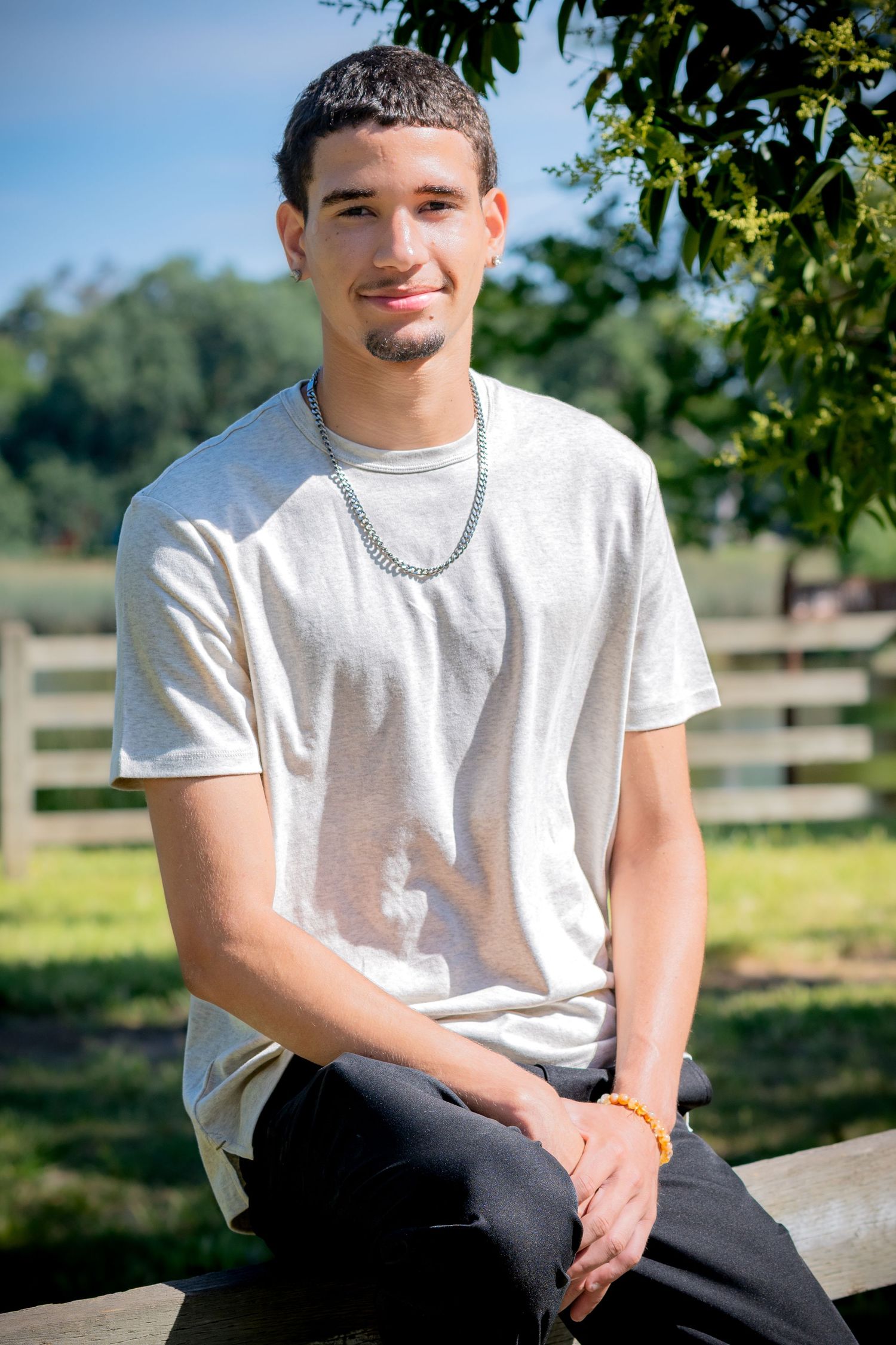 Young man in white t-shirt and chain necklace sitting on fence in sunny outdoor park setting.