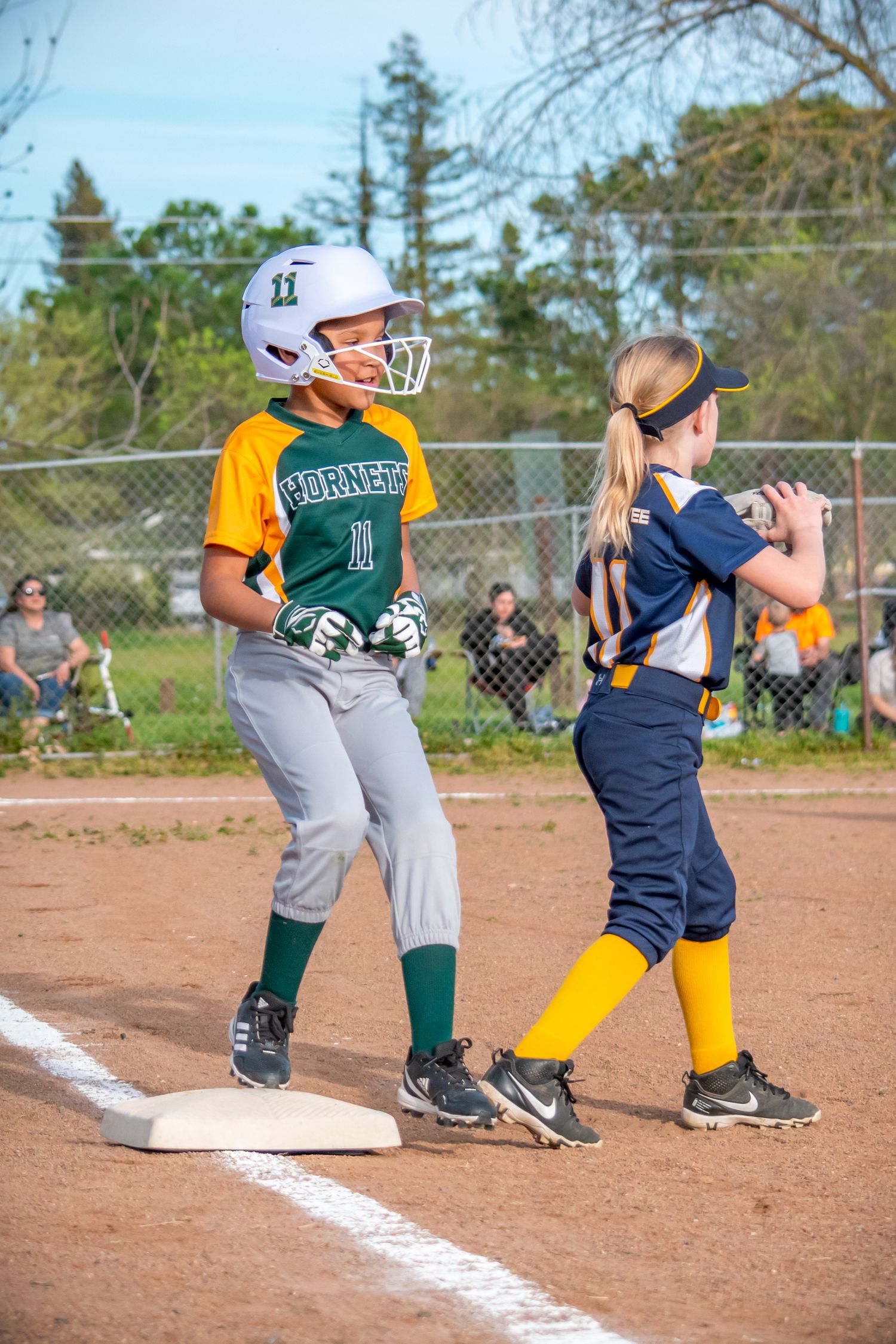 Youth Softball players running bases in green and yellow uniforms during a game on a dirt field.