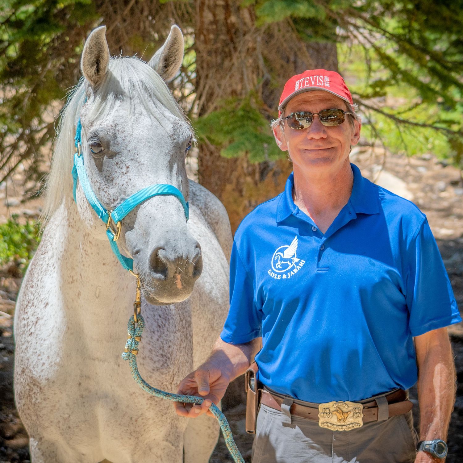 A light-colored horse stands next to someone wearing a bright blue polo shirt in a wooded area.