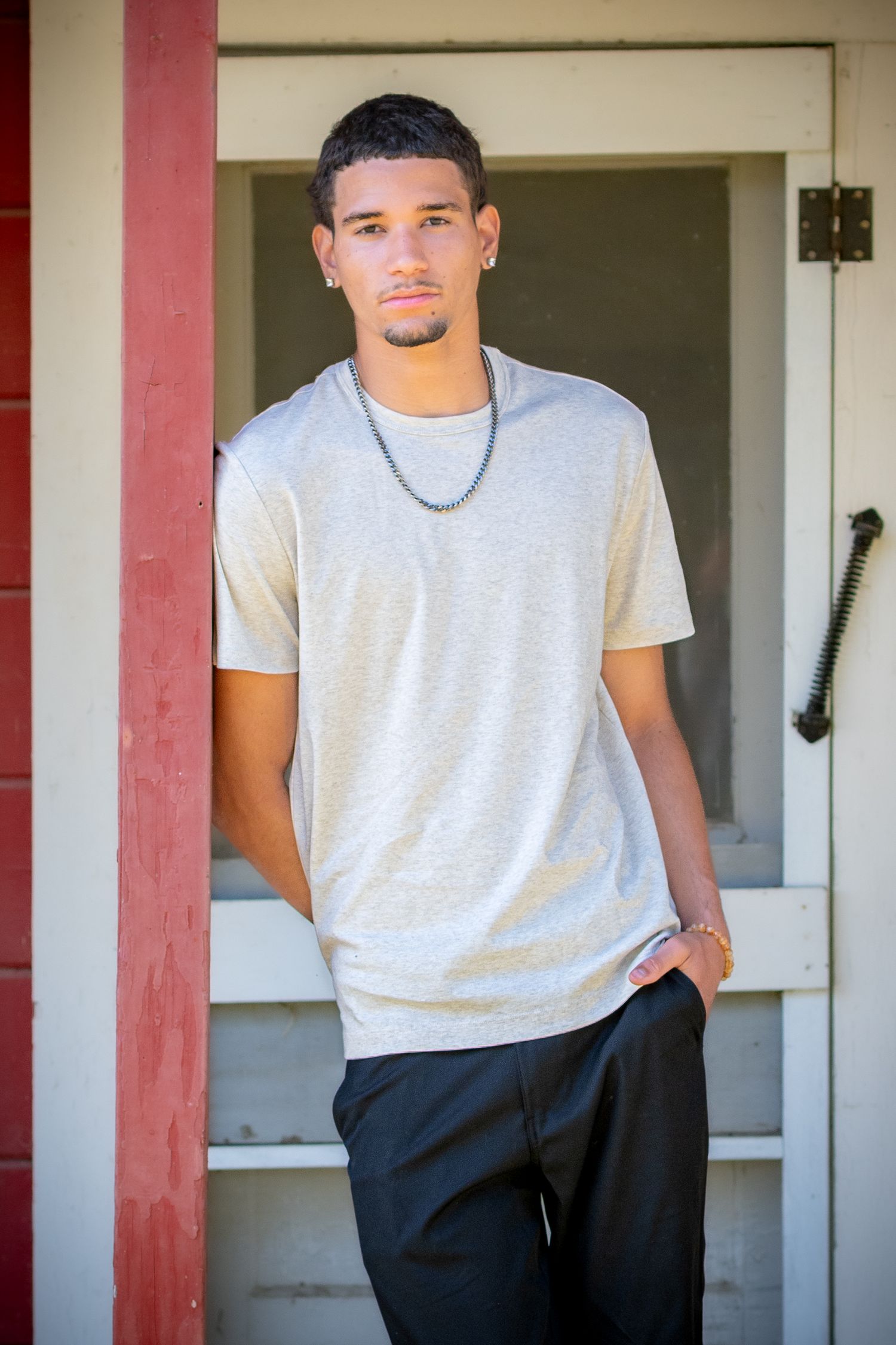 Young man in gray t-shirt and black pants leaning against a red wooden door frame outdoors.
