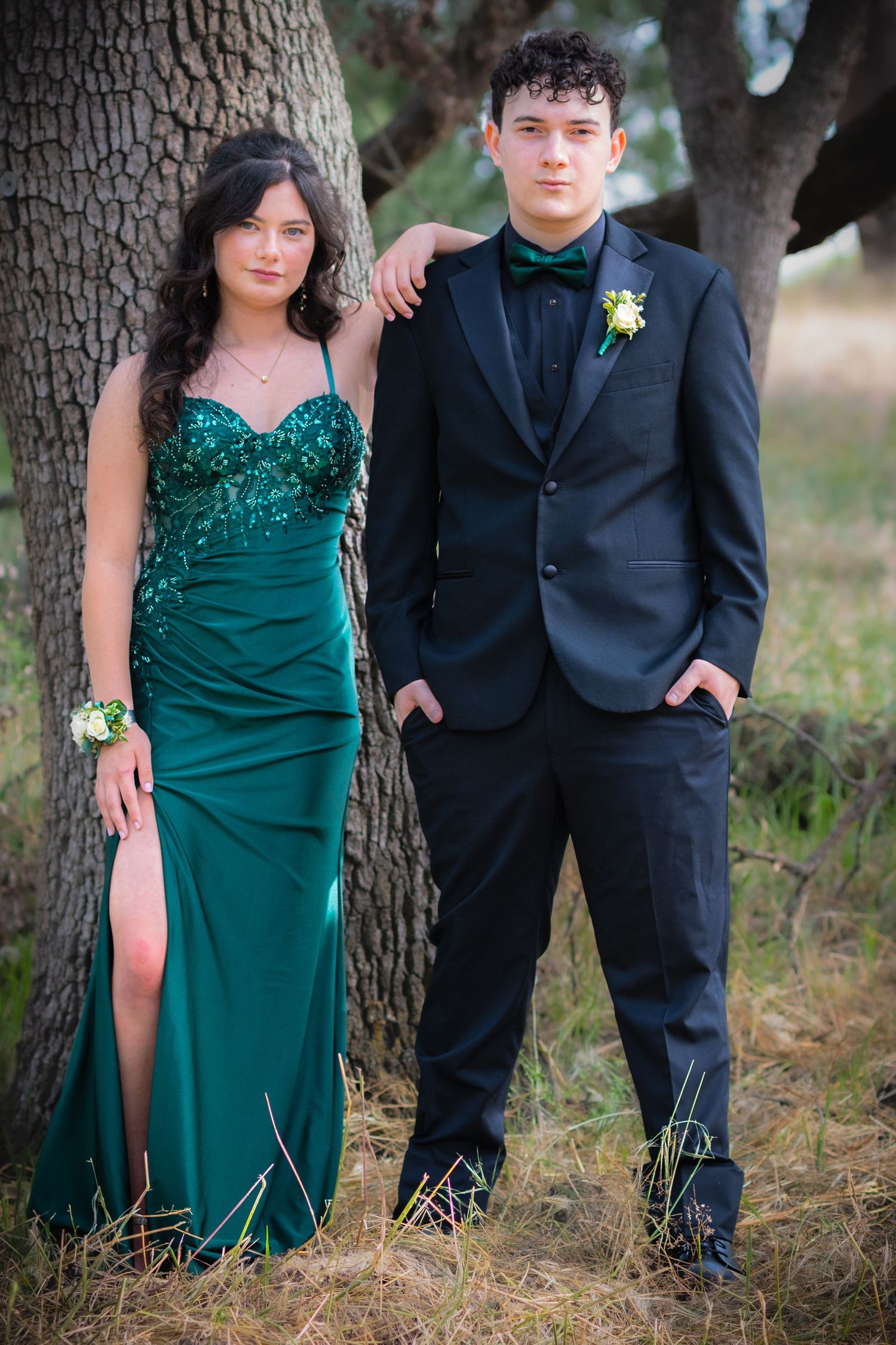 Teen couple in matching emerald green prom attire poses outdoors, girl in sequined gown, boy in black tuxedo with bow tie.