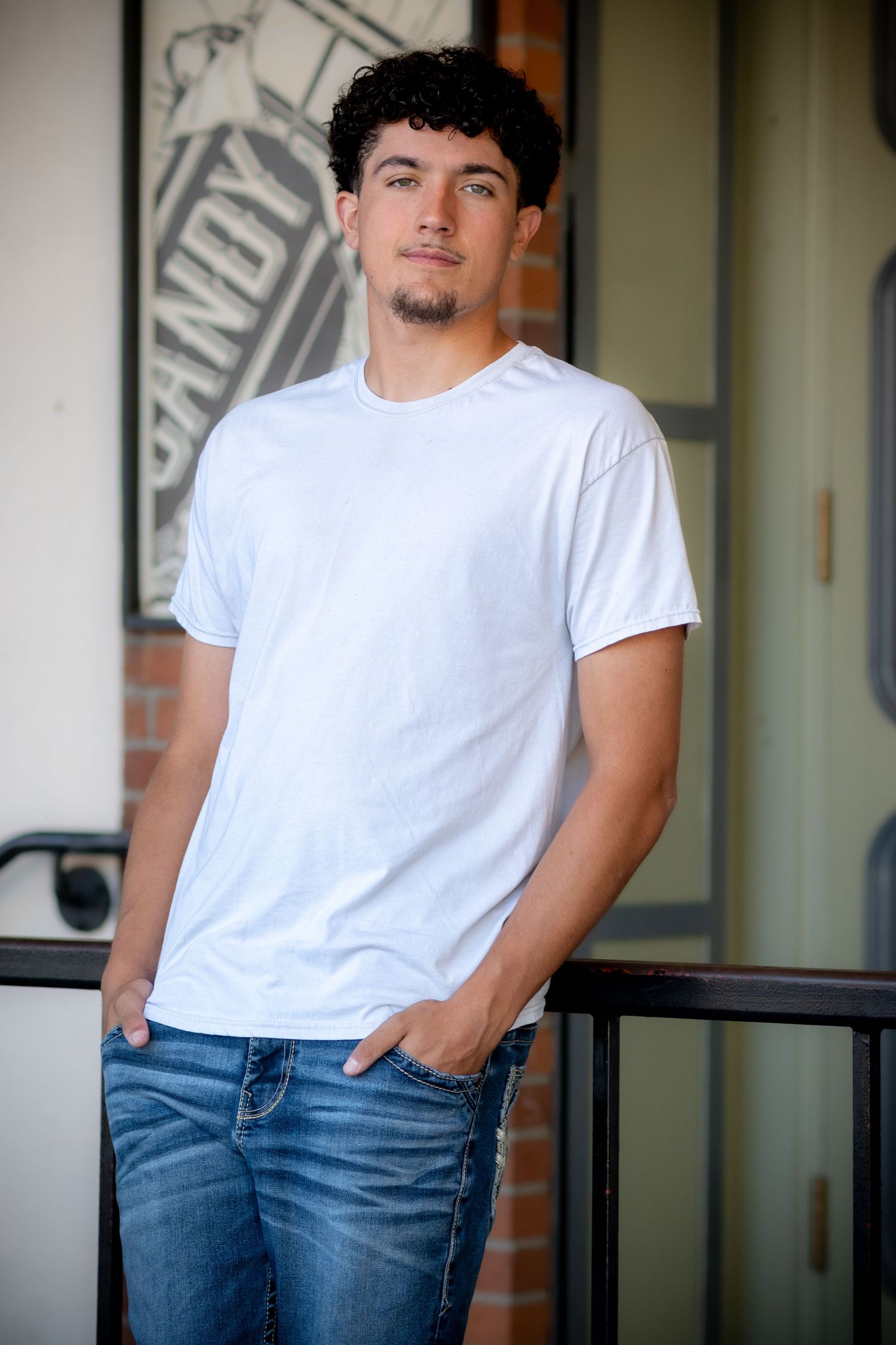 Young man with curly hair and goatee wearing white t-shirt and jeans, standing confidently outdoors.
