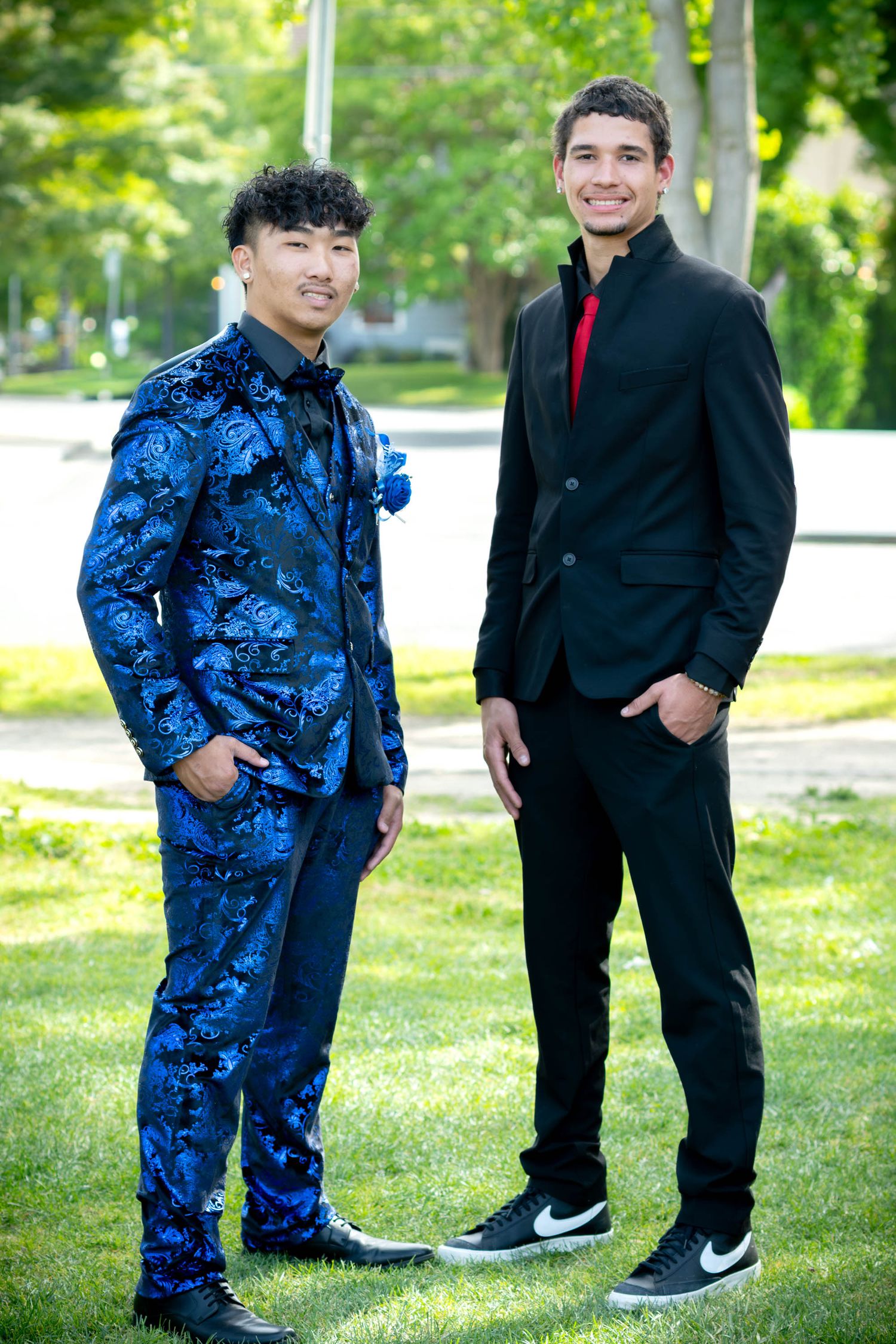 Two young men in formal suits posing outdoors on green grass for prom photos.