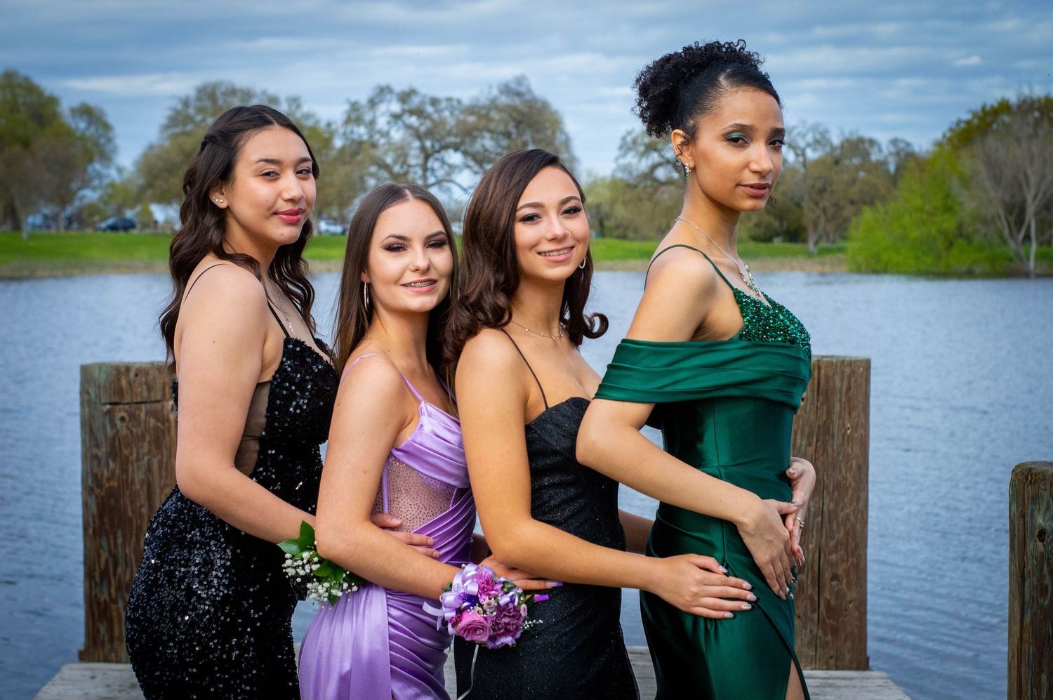 Four young women in elegant prom dresses pose together on a dock by a lake.