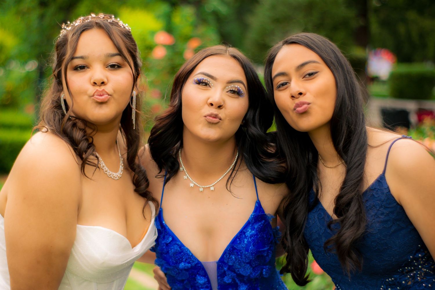 Three friends in formal dresses make playful expressions in a garden setting.