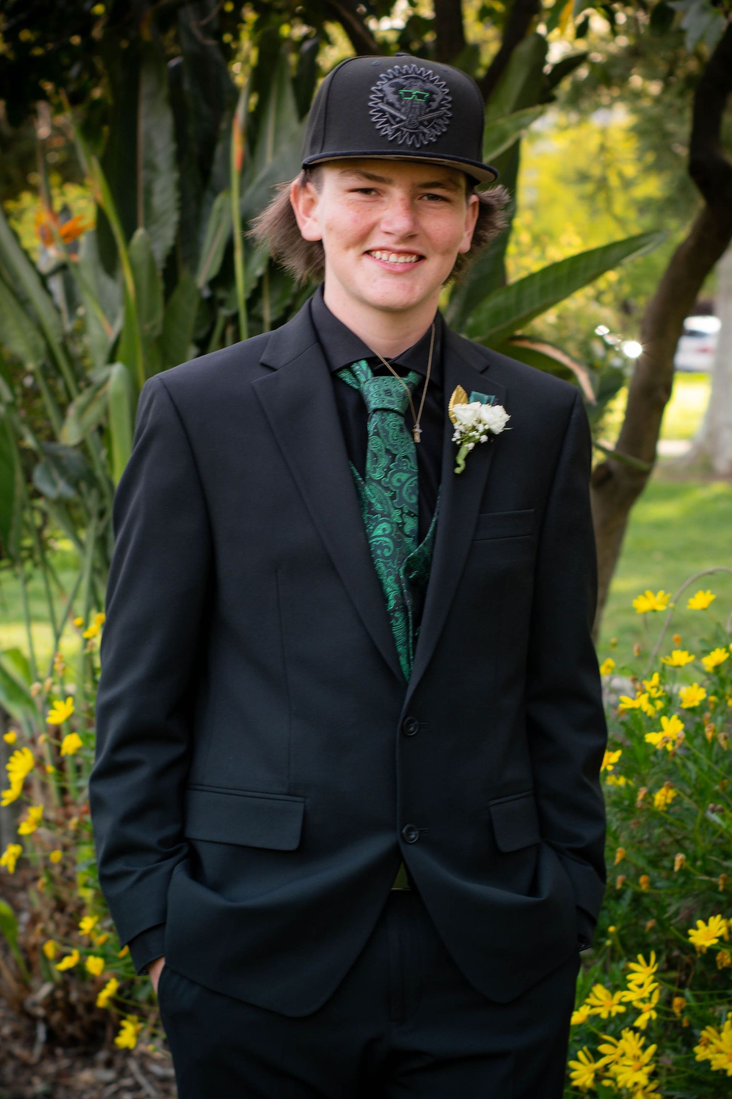 A person in a black suit with green vest and baseball cap poses in front of yellow flowers.