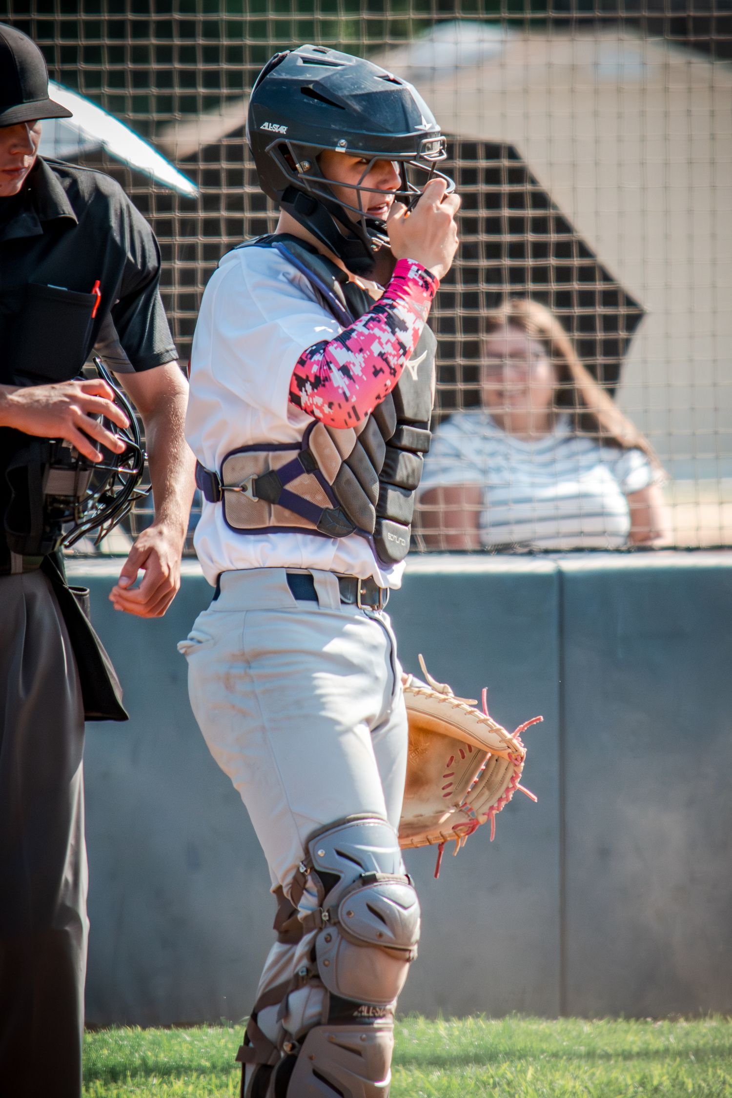 Catcher in baseball uniform with pink arm sleeve signals during game beside umpire on baseball field.