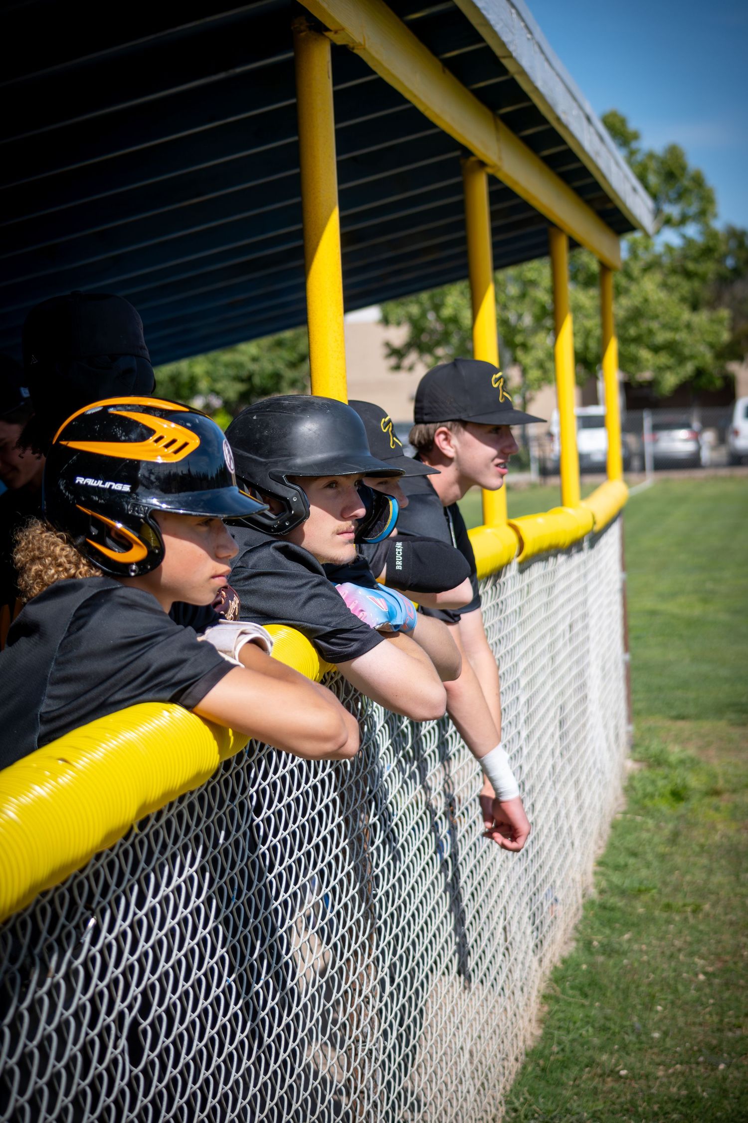 Baseball players in black uniforms watch the game from the dugout behind a chain-link fence with yellow rails.