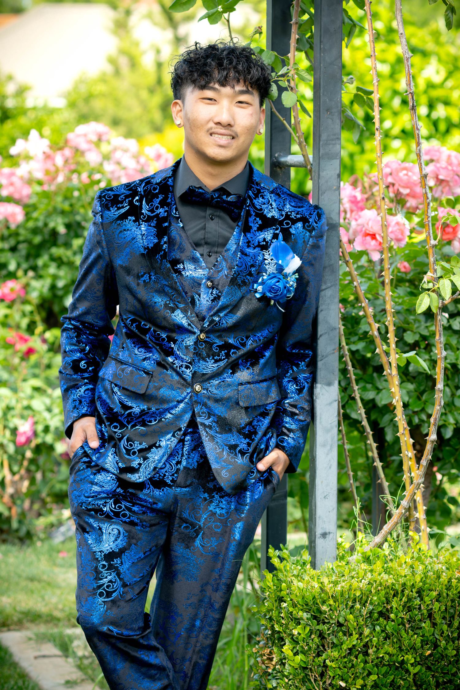 Young man in blue floral velvet tuxedo poses in garden with pink roses.