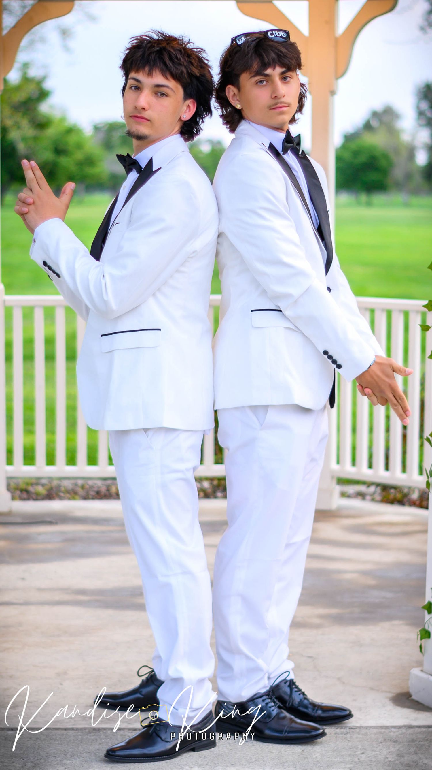 Two young men in matching white tuxedos with black lapels pose back-to-back outdoors near a gazebo.