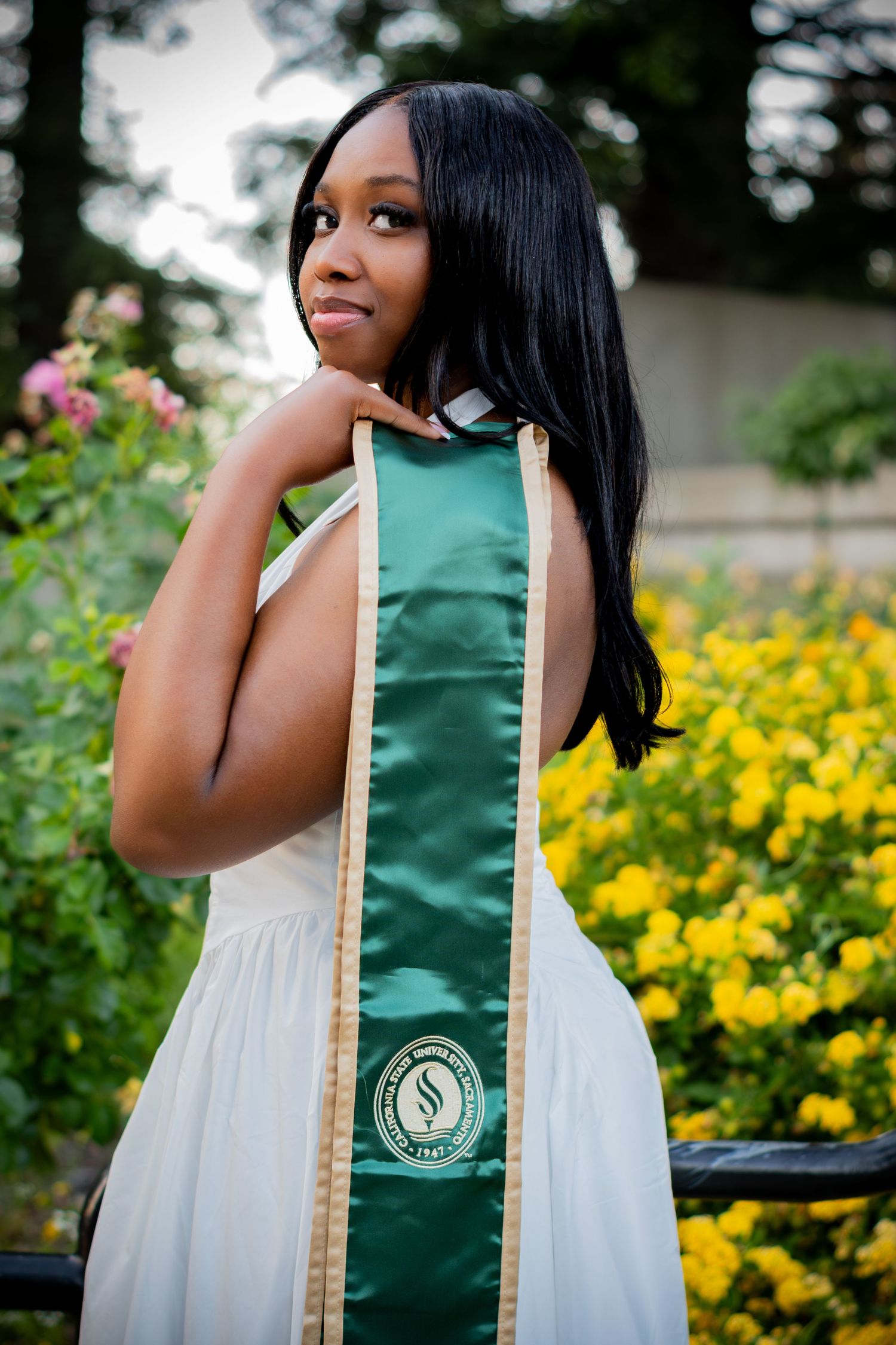 Young woman in white dress wearing green graduation stole poses outdoors among yellow flowers.