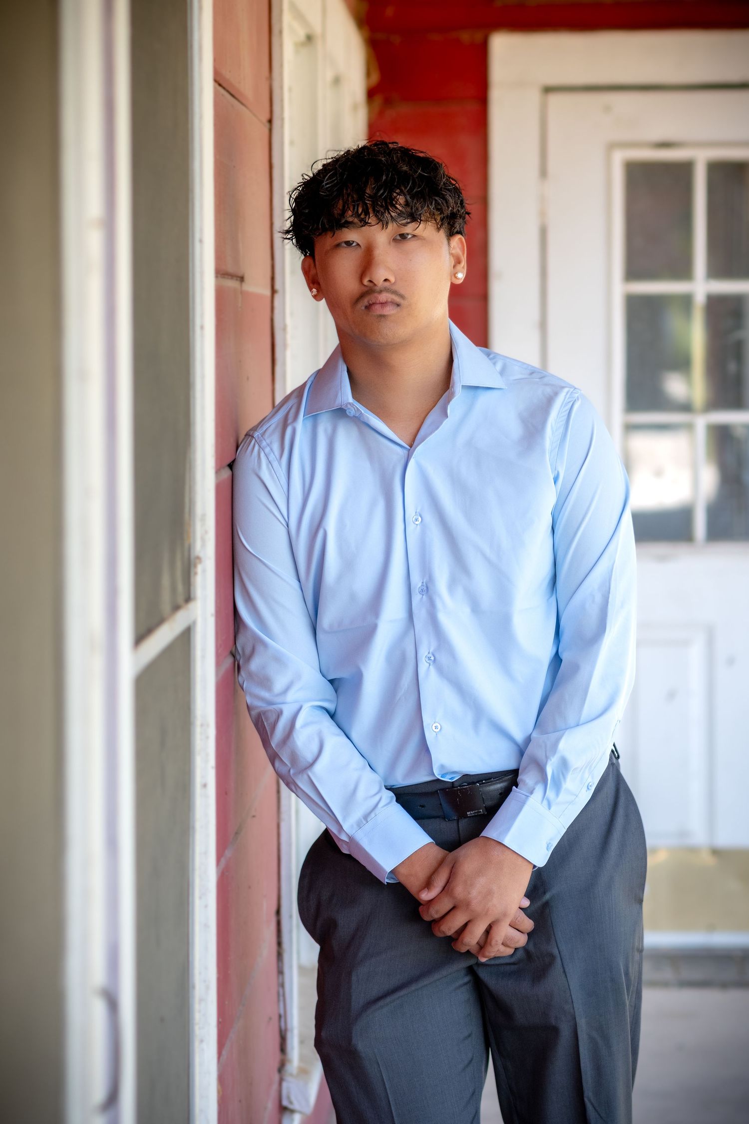 Young man in light blue dress shirt and gray trousers leaning against a red building exterior.