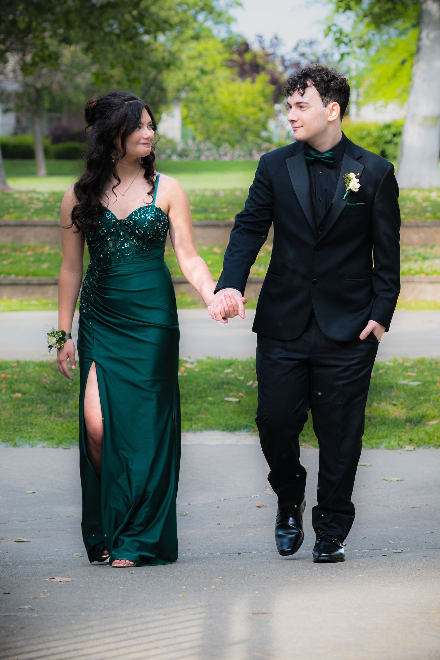 A couple in formal prom attire walks hand-in-hand, she in emerald green gown, he in black tuxedo, outdoors.