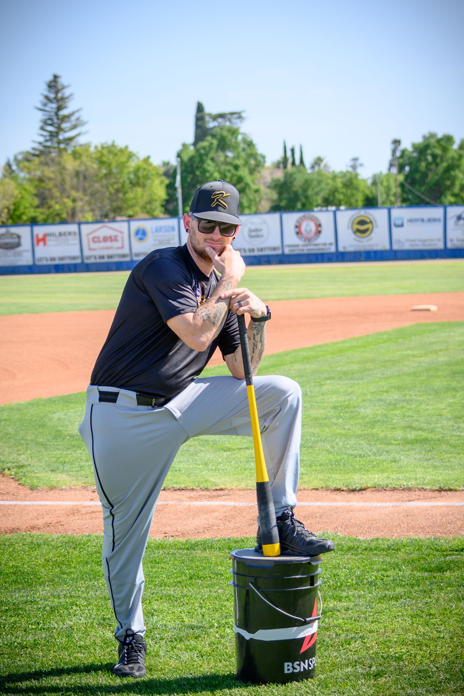 Baseball coach sitting on equipment bucket dugout on baseball field during sunny day.