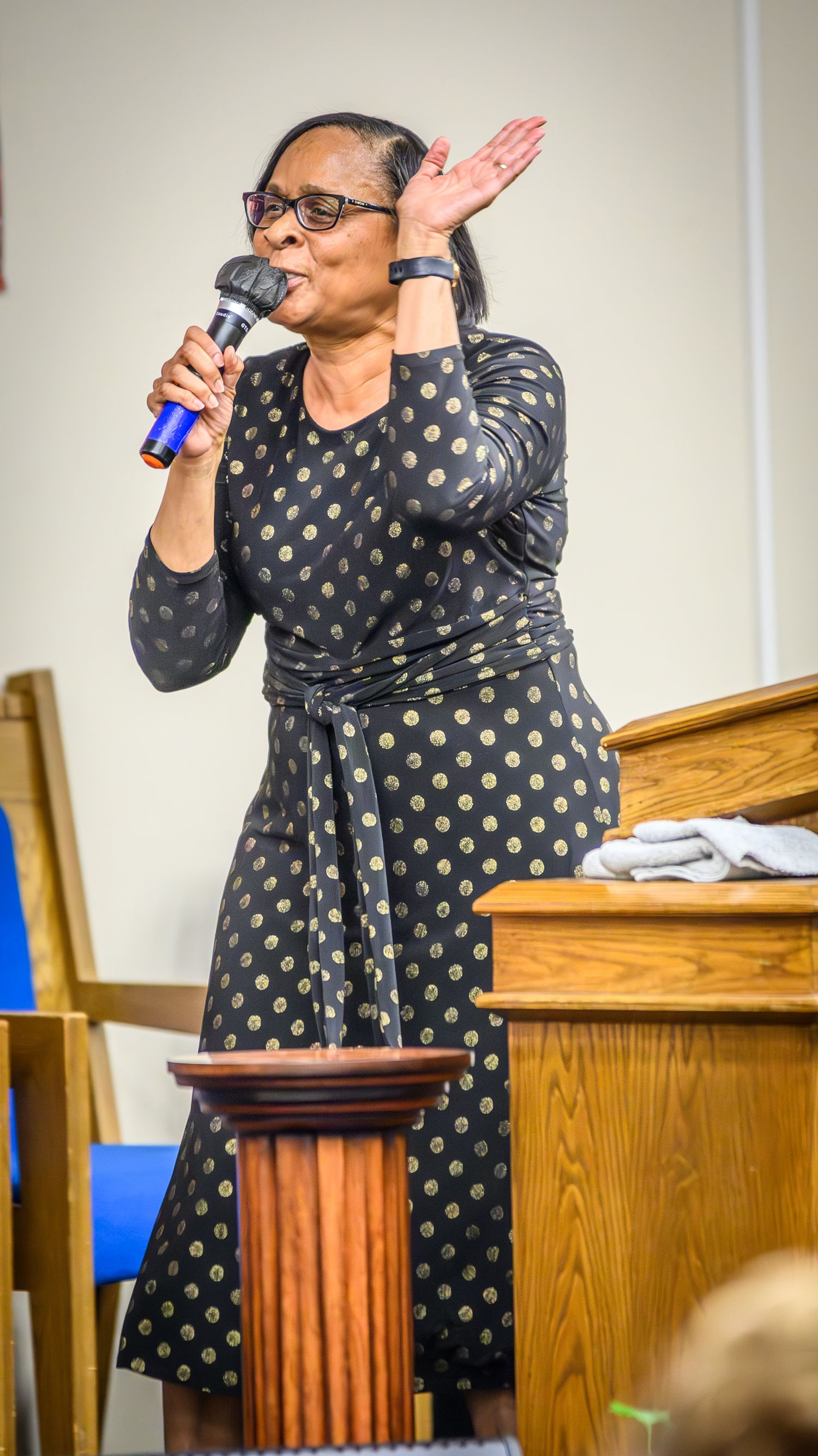 A speaker in a polka dot wrap dress delivers an animated presentation at a wooden podium.