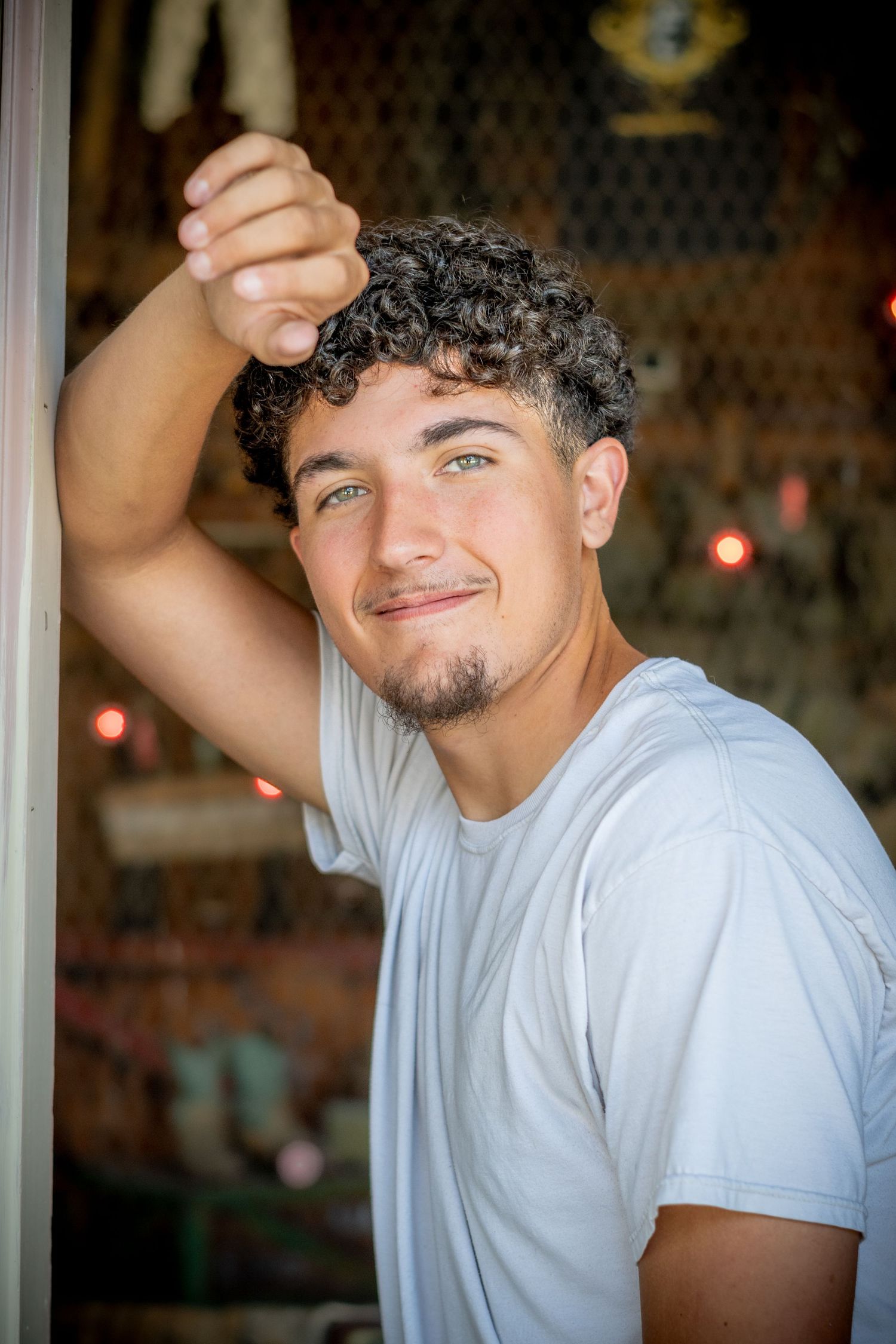 Young man with curly hair and green eyes smiling in a white t-shirt, leaning against a doorframe.