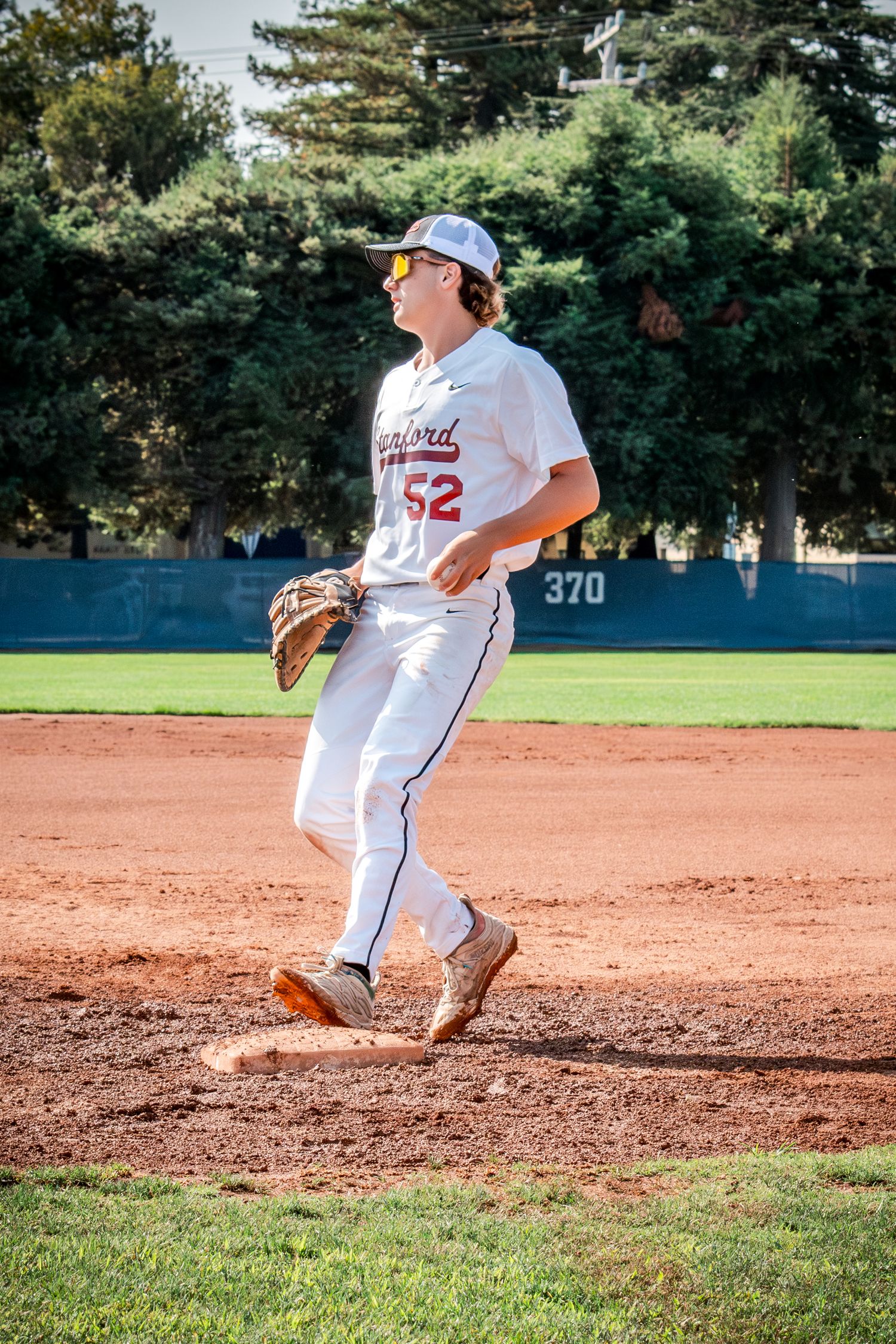 Baseball player slides into base during game on dirt infield with trees visible in background.