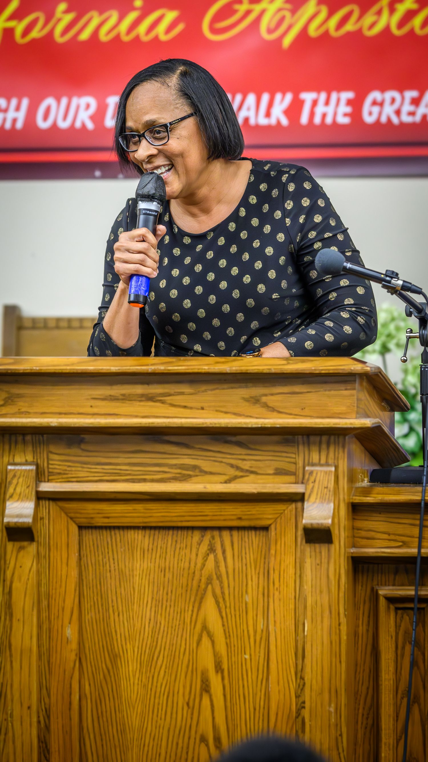 A speaker in a polka dot dress stands at a wooden podium smiling and gesturing during a presentation.