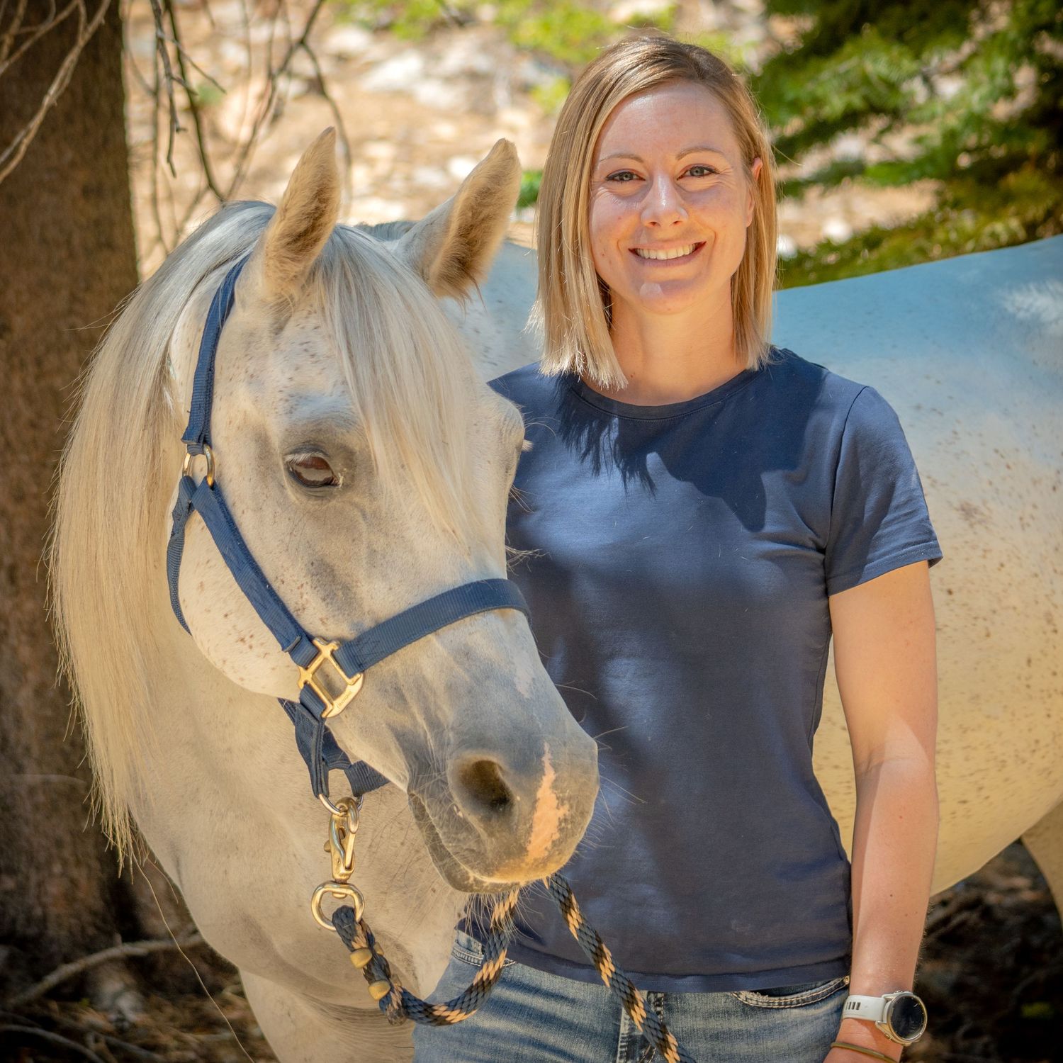 A white horse with blue halter poses with someone in a navy shirt against a natural background.