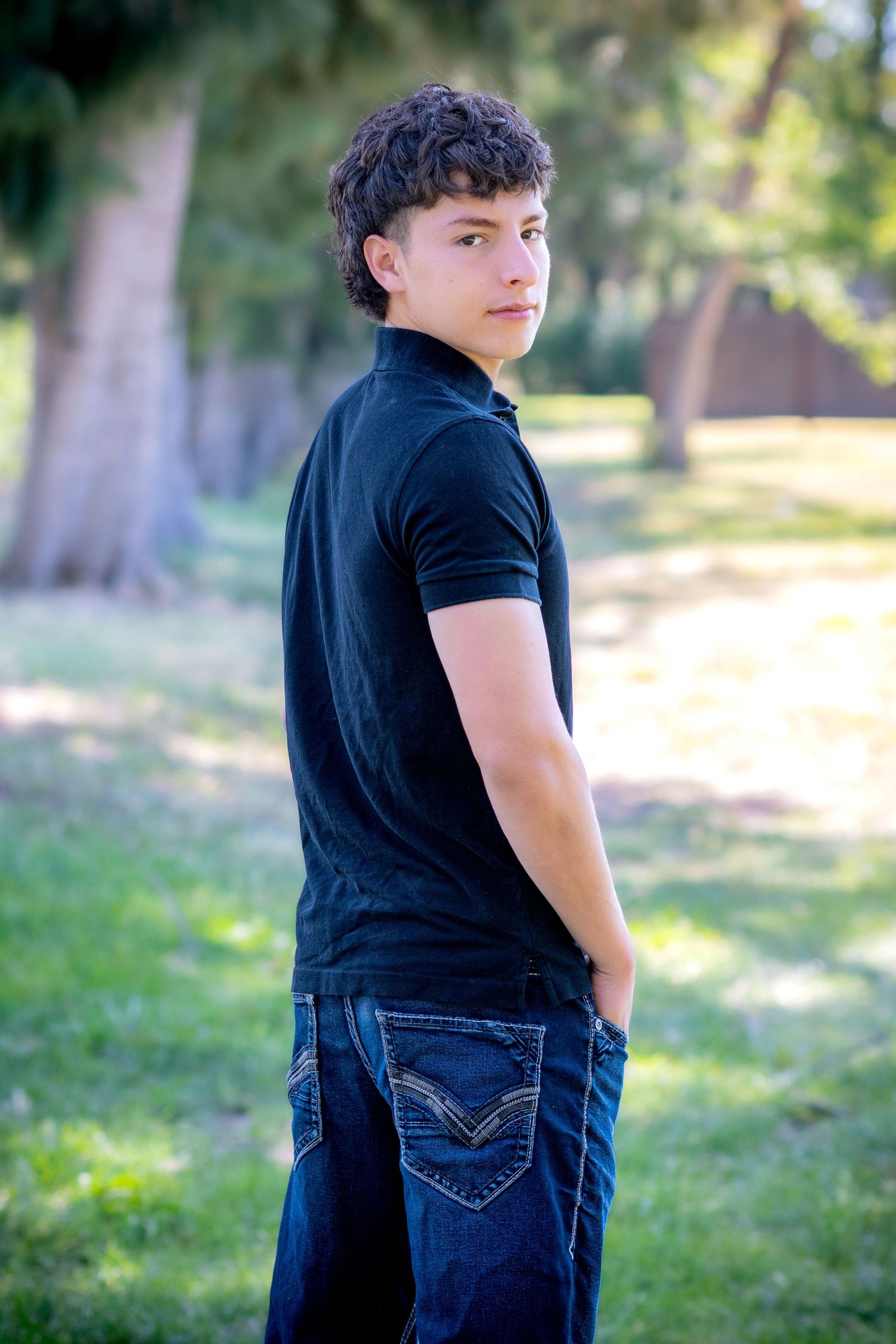 Teen boy in black polo shirt and jeans stands in a park, looking back over his shoulder with a confident expression.