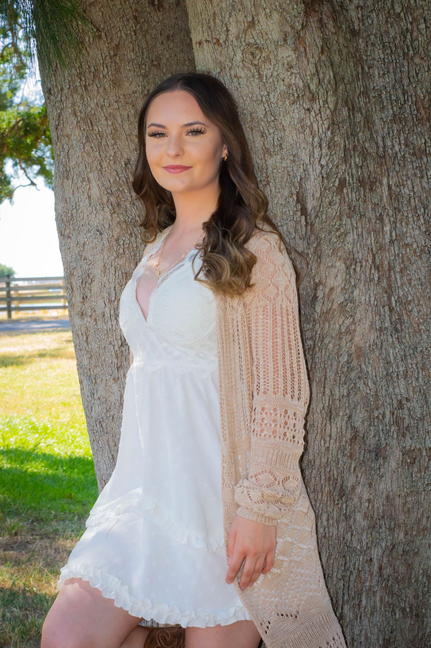 Person in white lace dress and beige cardigan poses by large tree trunk in sunny outdoor setting.