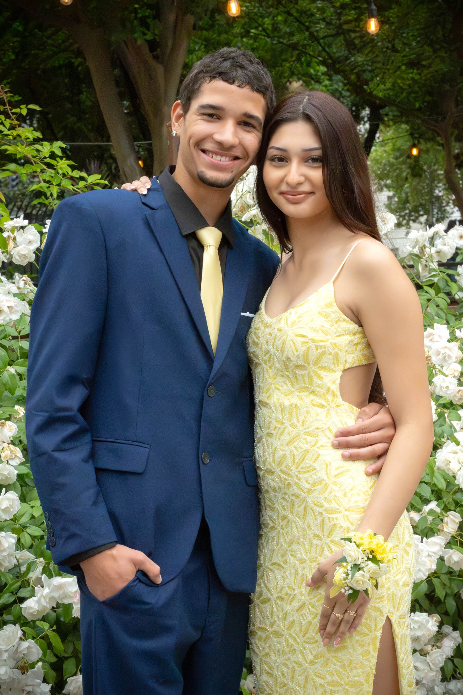 A smiling couple dressed in prom attire, man in navy suit and woman in yellow dress, pose among white flowers.