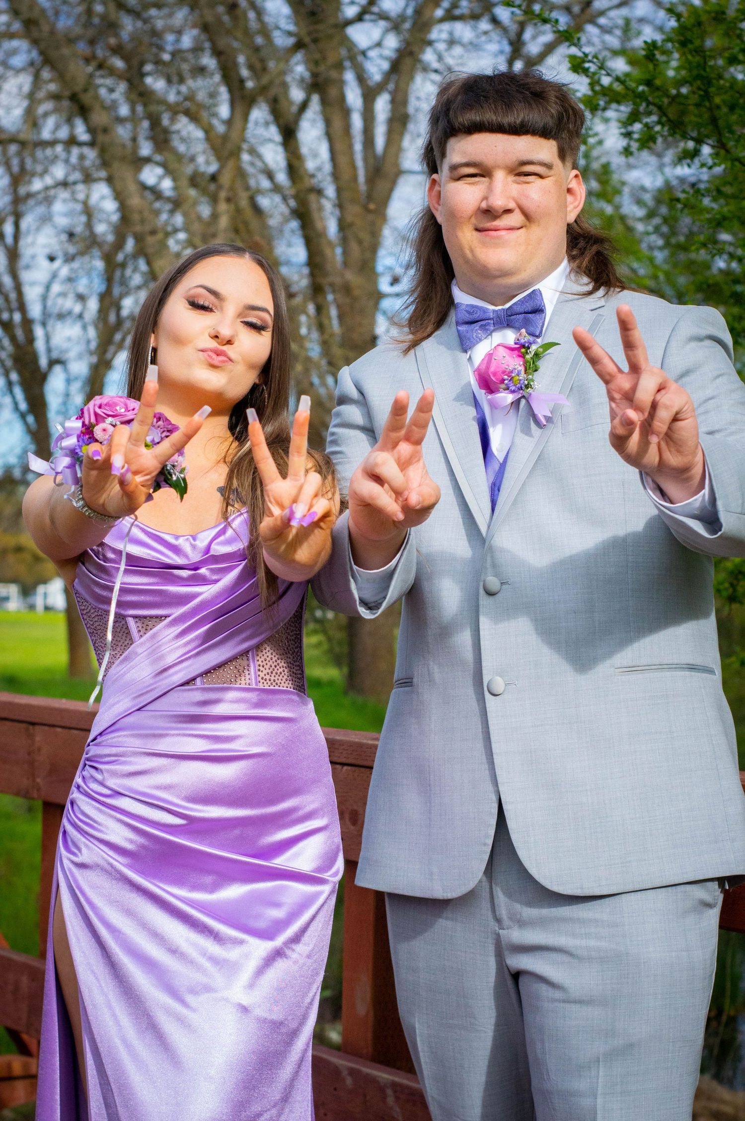 A couple in purple dress and grey suit make playful peace signs while posing outdoors in a park setting.