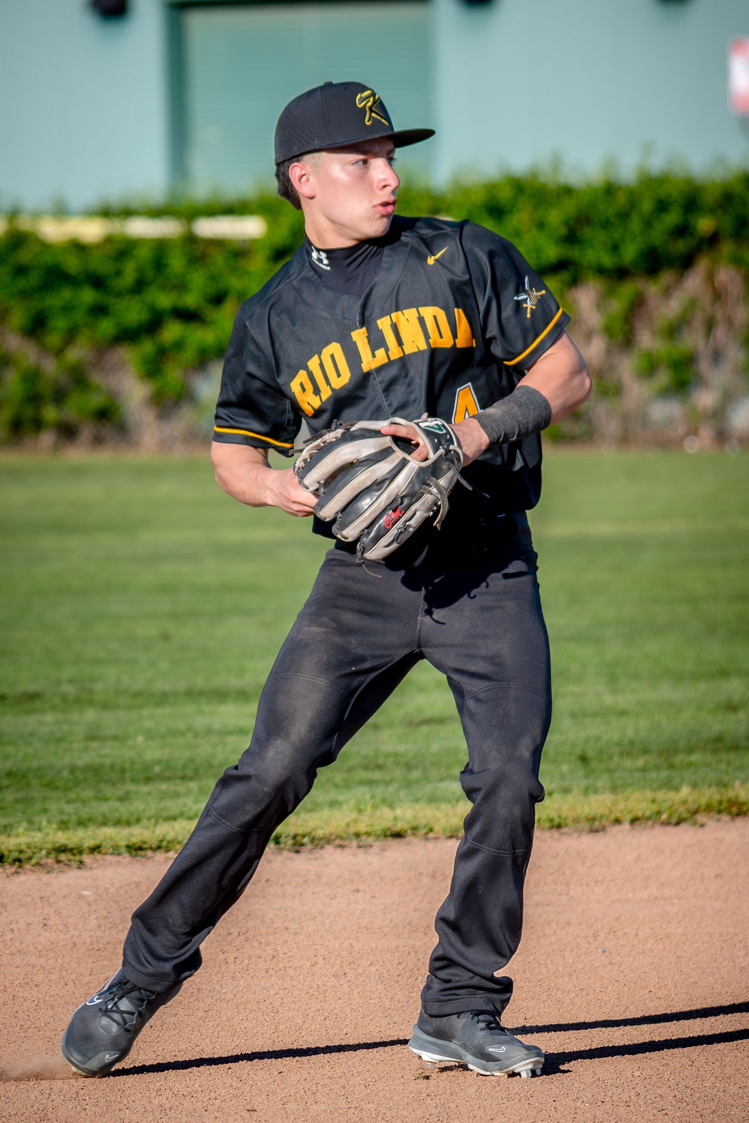 Rio Linda baseball player in black uniform fielding position on dirt infield, wearing glove and cap.
