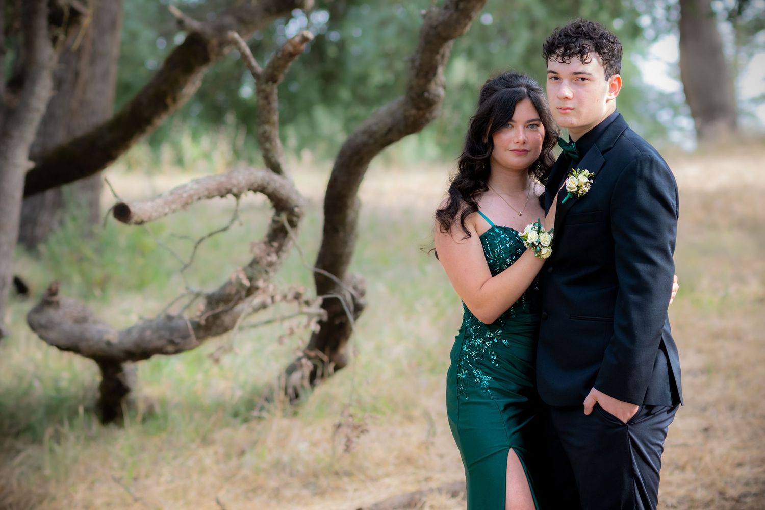 A couple in formal prom attire poses outdoors, she in a green sequin gown, he in a black suit.