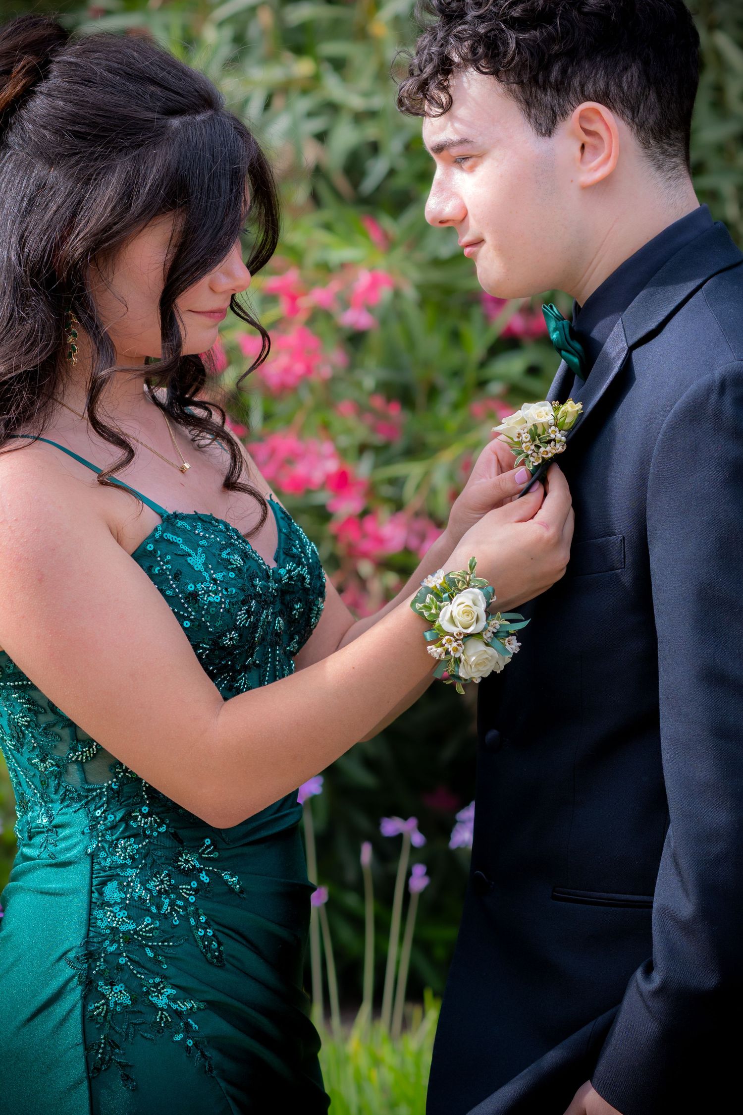 Teen couple in prom attire, girl in green gown pins boutonniere on boy in black suit outdoors.