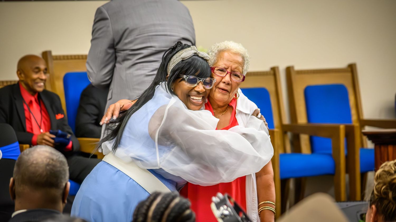 People seated in blue chairs participate in an indoor community gathering or service.