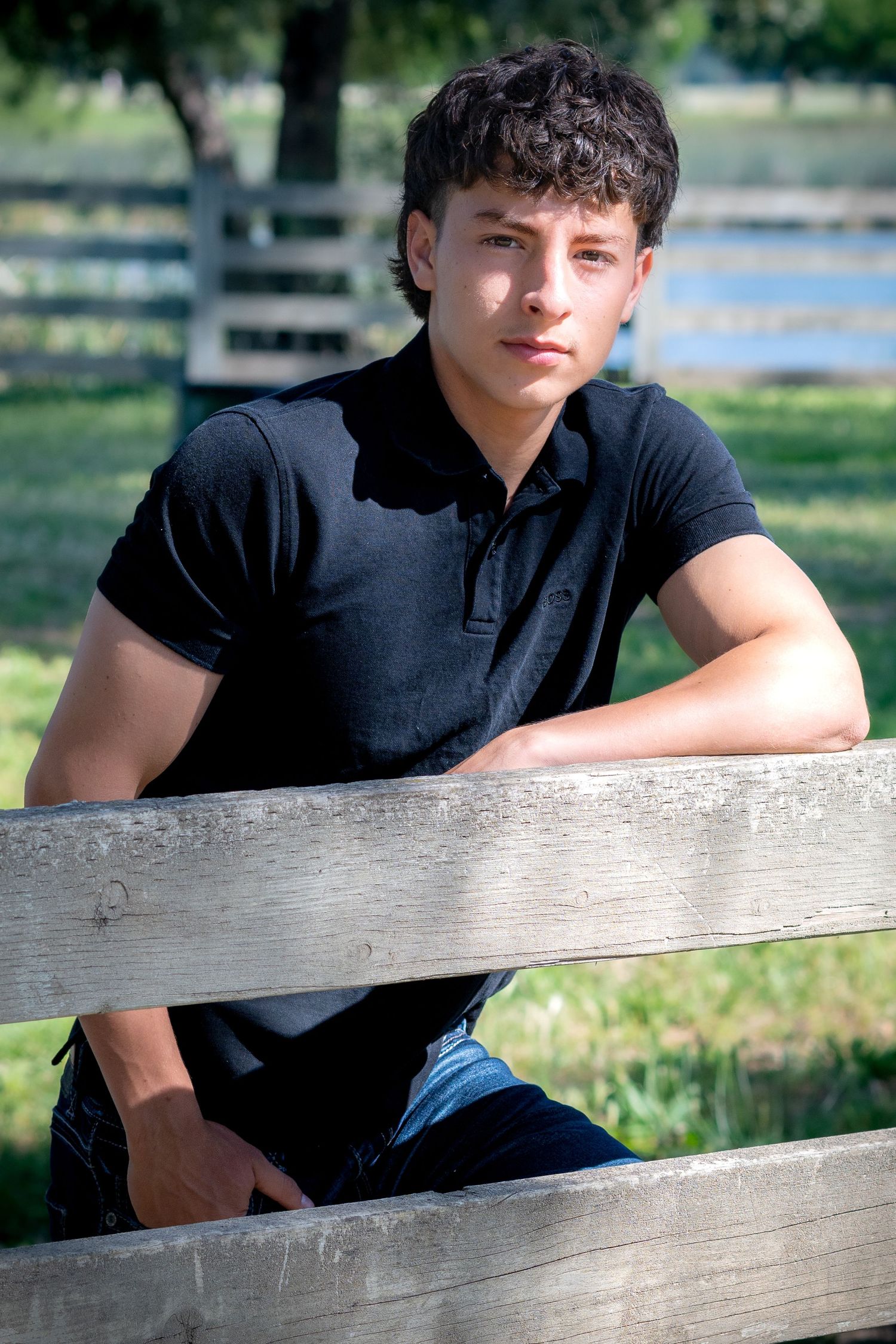 Young man in black polo shirt leaning on wooden fence in rural setting, looking at camera.
